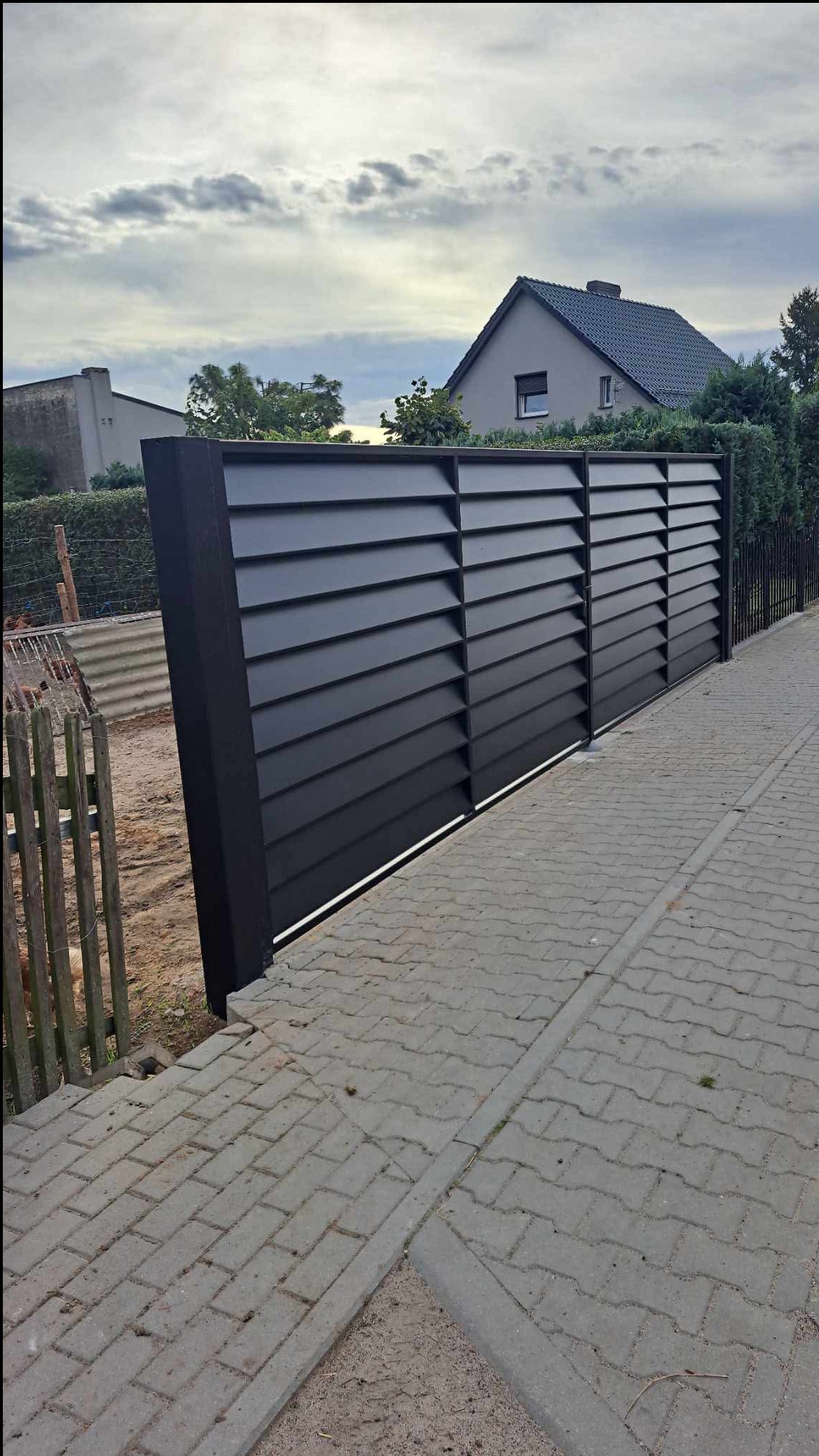 Nowoczesne, poziome, czarne panele ogrodzeniowe z metalu, zamontowane na posesji. Widok z boku, z kostką brukową i fragmentem starego, drewnianego płotu.