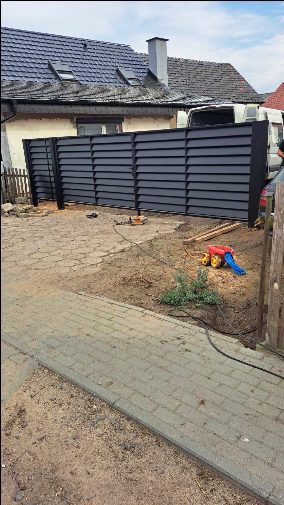Nowoczesne, poziome ogrodzenie panelowe w kolorze antracytowym, częściowo zamontowane. Widoczne narzędzia i zabawka dziecka na posesji. Dom z szarą dachówką w tle.