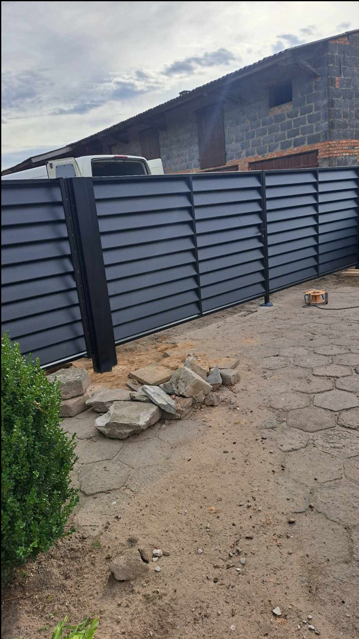 Nowoczesne, poziome przęsła ogrodzeniowe w kolorze antracytu, z widocznymi elementami montażowymi i stertą kamieni u podstawy. W tle budynek gospodarczy i fragment samochodu.