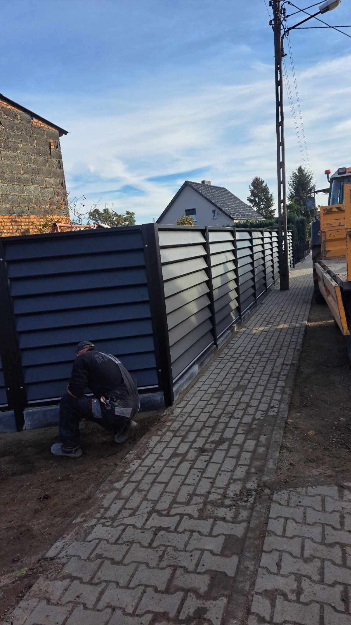 Montaż nowoczesnego, poziomego ogrodzenia panelowego w kolorze grafitowym. Widoczny pracownik w trakcie instalacji, kostka brukowa i budynki w tle.