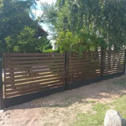 Nowoczesne ogrodzenie posesji z poziomych, brązowych paneli na czarnej podmurówce. Widoczne drzewa i trawa.