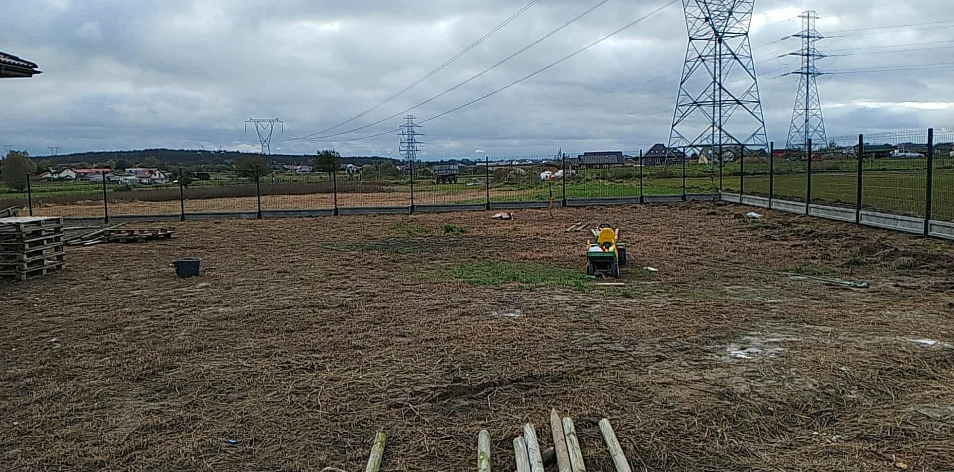 Montaż ogrodzenia panelowego z betonową podmurówką na posesji, widok na teren przygotowany pod trawnik, w tle słupy wysokiego napięcia i zabudowa wiejska.