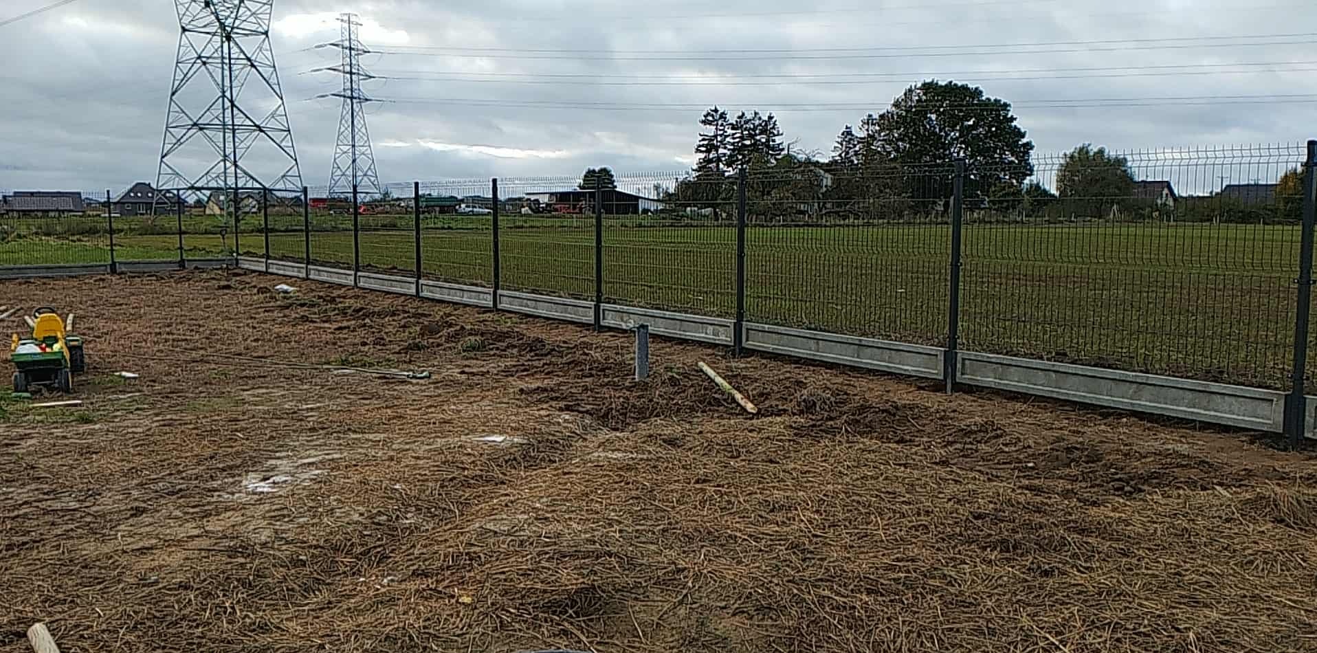 Nowoczesne ogrodzenie panelowe z podmurówką betonową, widok na rozległy teren z liniami wysokiego napięcia w tle. Częściowo przygotowany teren pod dalsze prace.