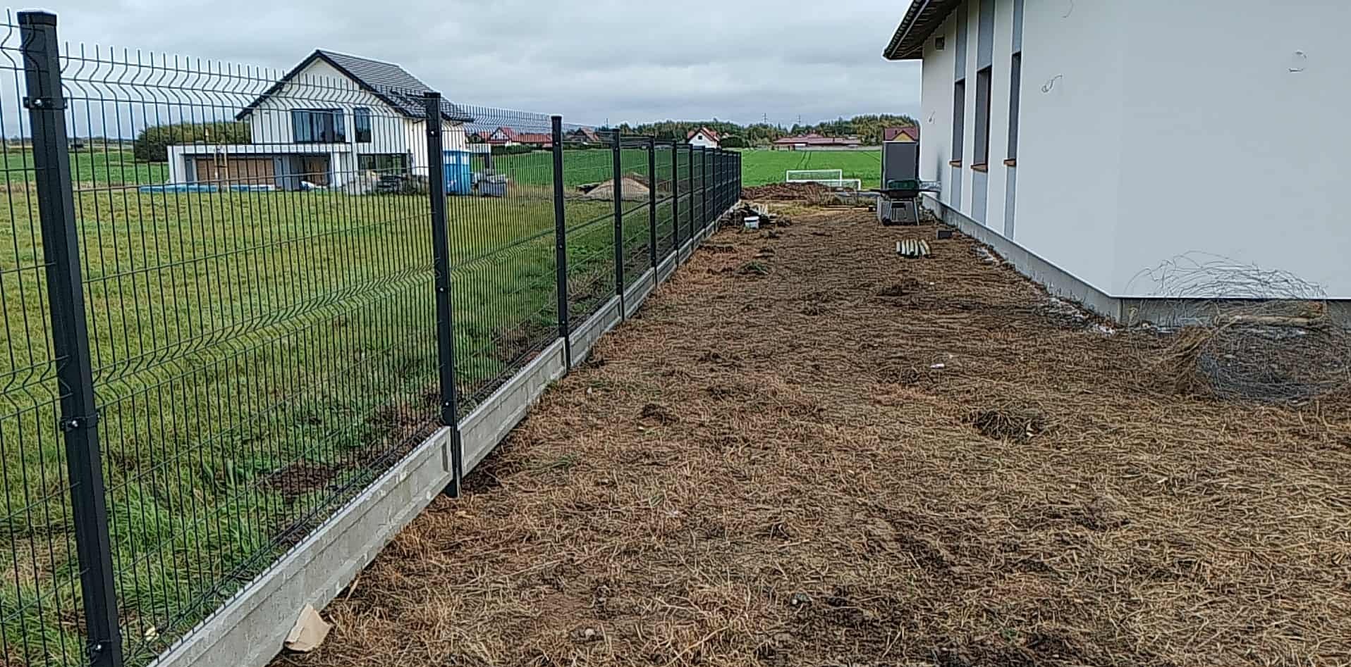 Nowoczesne ogrodzenie panelowe z podmurówką betonową, widok z boku posesji, w tle dom jednorodzinny i krajobraz wiejski pod pochmurnym niebem.