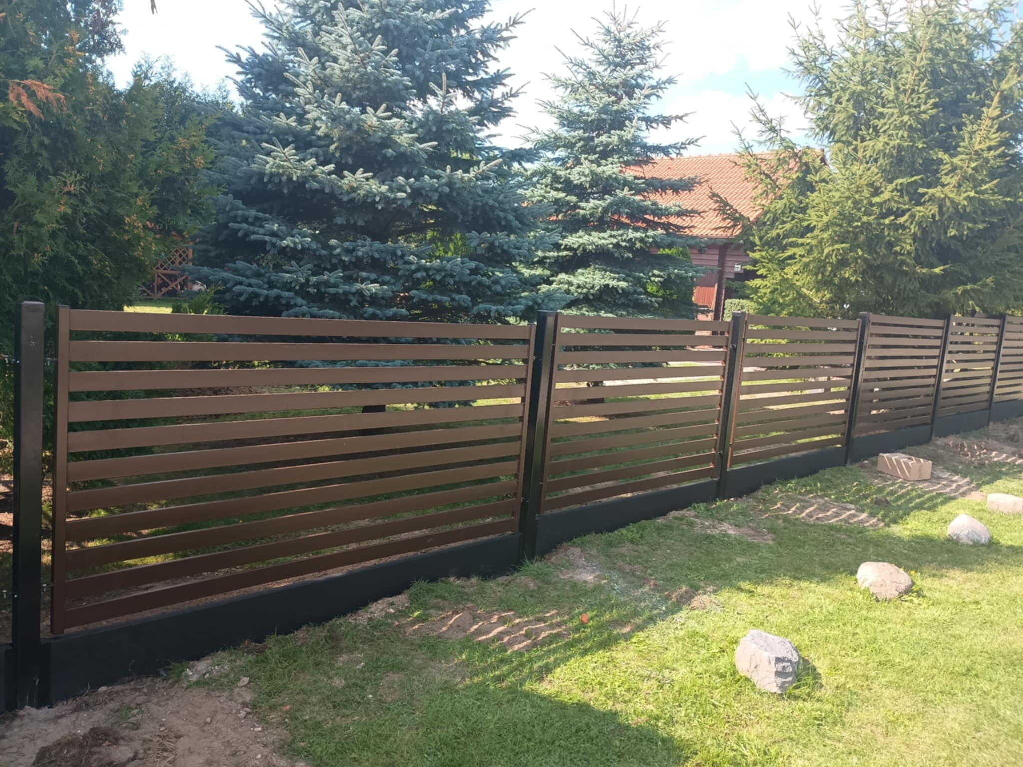 Nowoczesne ogrodzenie panelowe z poziomych listew w kolorze brązowym, na tle zieleni drzew i trawnika, z widocznymi kamieniami dekoracyjnymi.