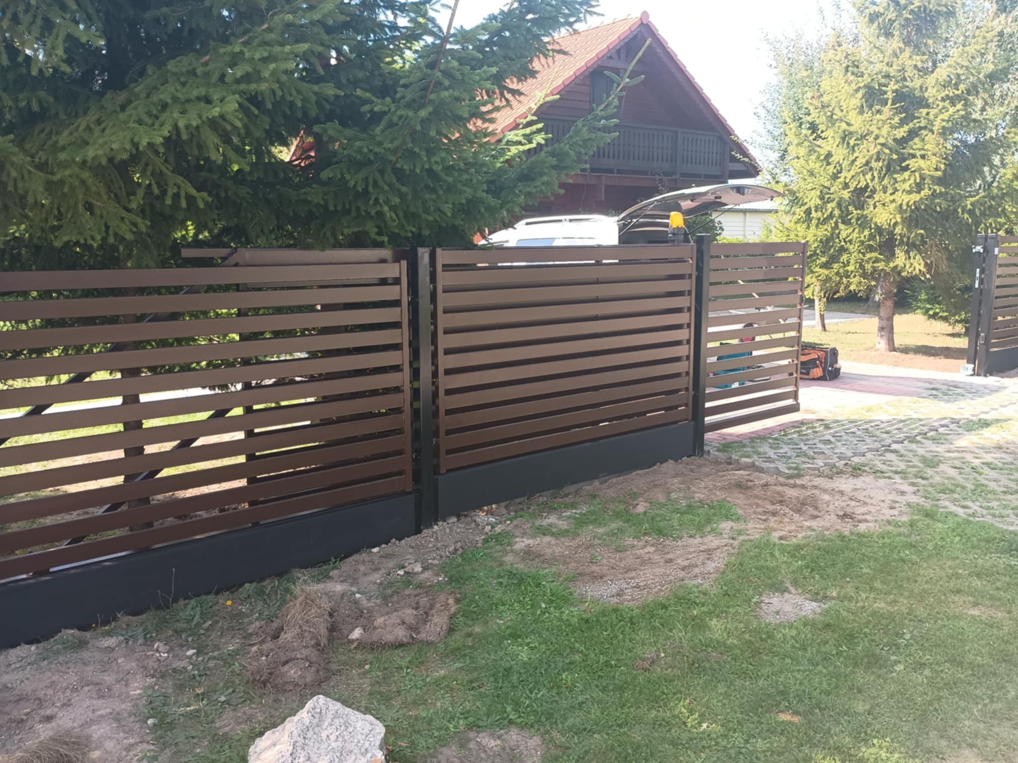 Nowoczesne ogrodzenie panelowe z poziomych, brązowych listew i czarną podmurówką,  na tle domu z czerwonym dachem i zieleni, częściowo otwarte wejście.