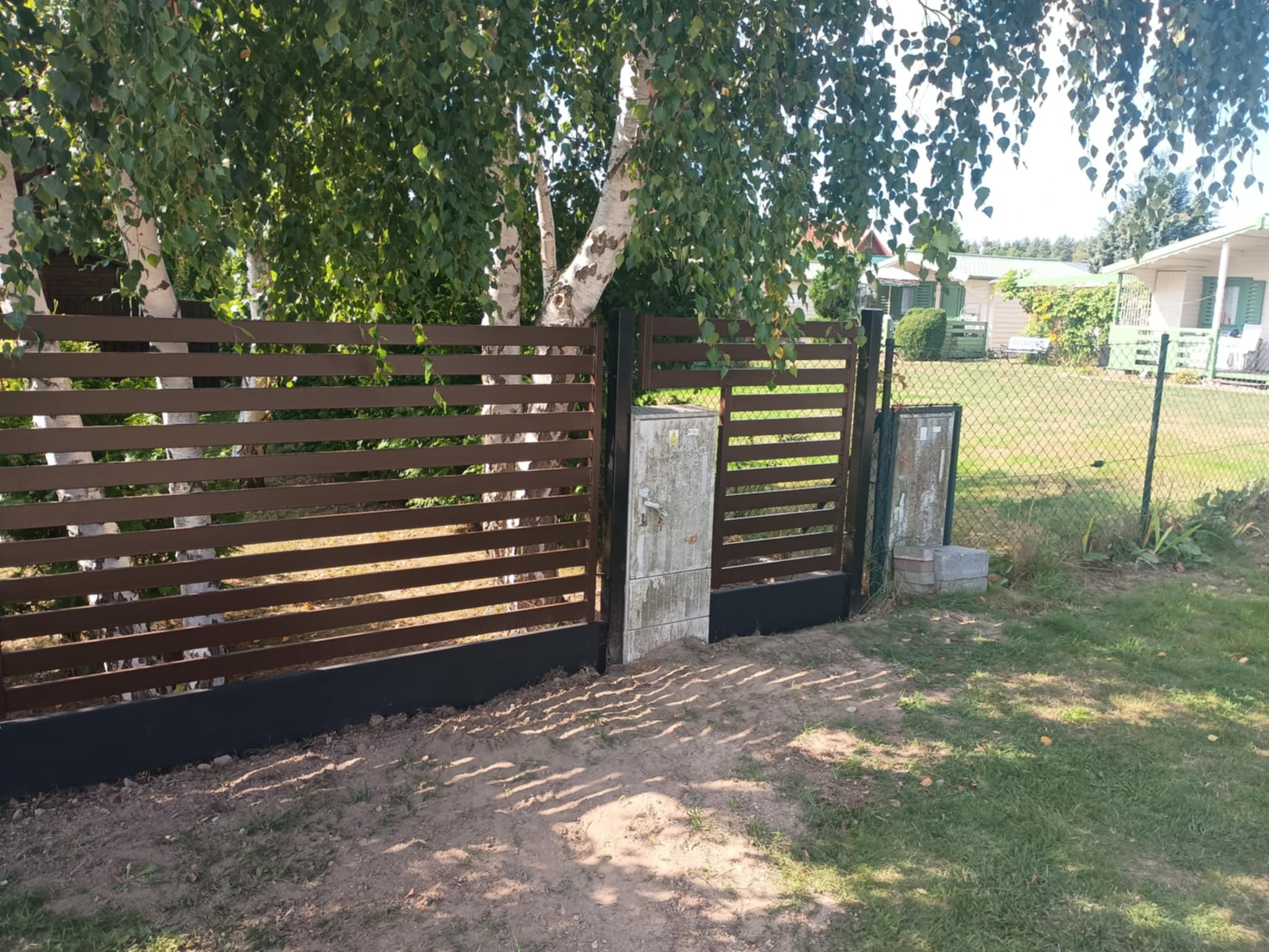 Nowoczesne, brązowe ogrodzenie panelowe z poziomych desek, z czarną podmurówką i słupkami, na tle zieleni drzew i zabudowań. Brama wjazdowa częściowo otwarta.