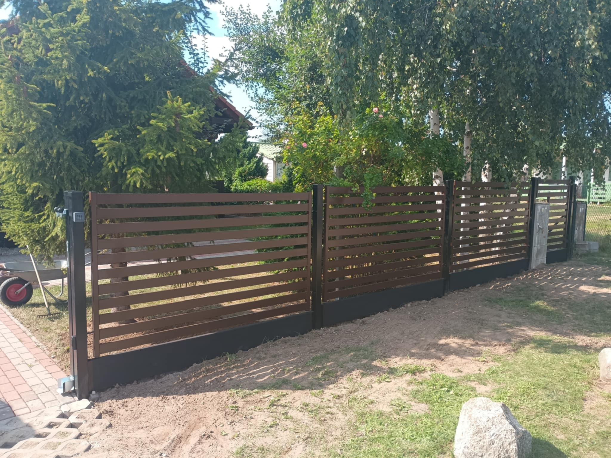 Nowoczesne ogrodzenie posesji z poziomych, brązowych paneli na czarnej podmurówce. Widoczne drzewa i trawa.