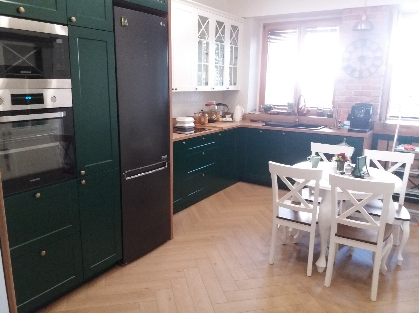 Nowoczesna kuchnia z ciemnozielonymi szafkami, białymi górnymi szafkami z przeszkleniami i parkietem ułożonym w jodełkę. Widoczny stół z krzesłami.