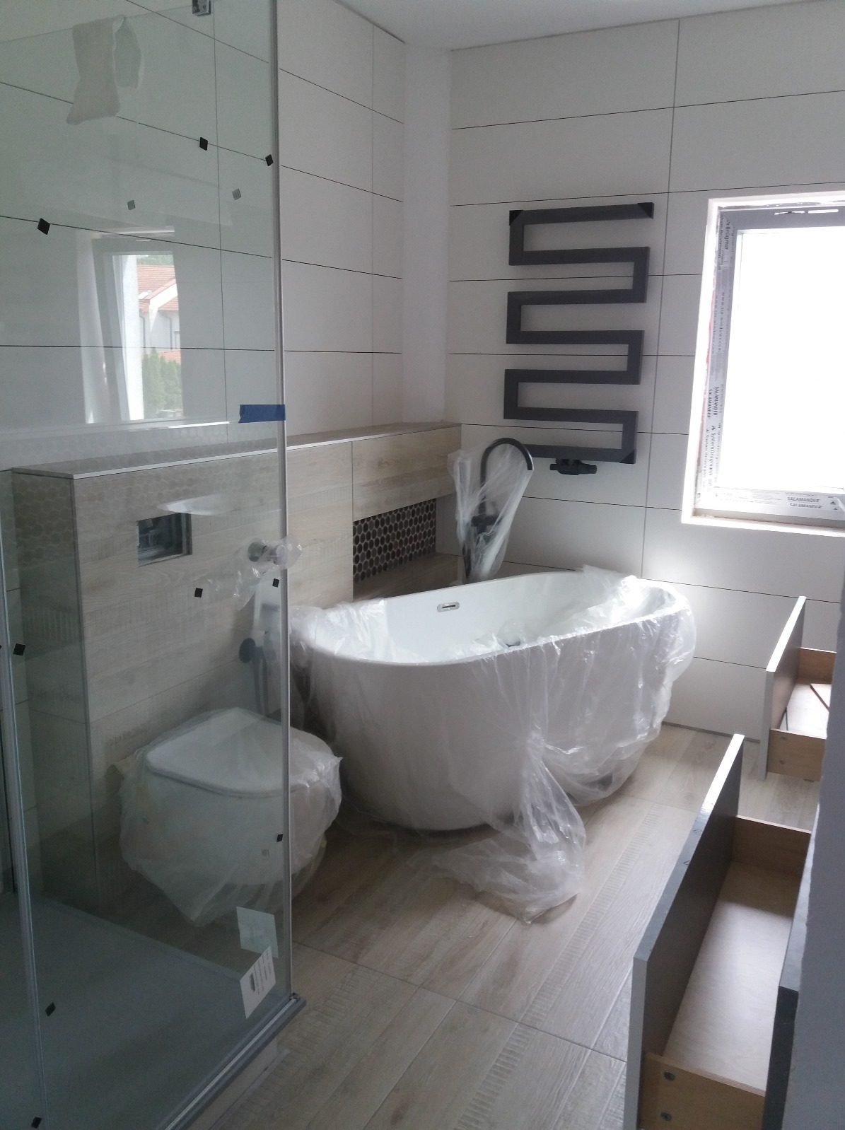 Nowoczesna łazienka z białą wanną wolnostojącą i szklaną kabiną prysznicową, jeszcze zabezpieczona folią ochronną. Na ścianie grzejnik drabinkowy.