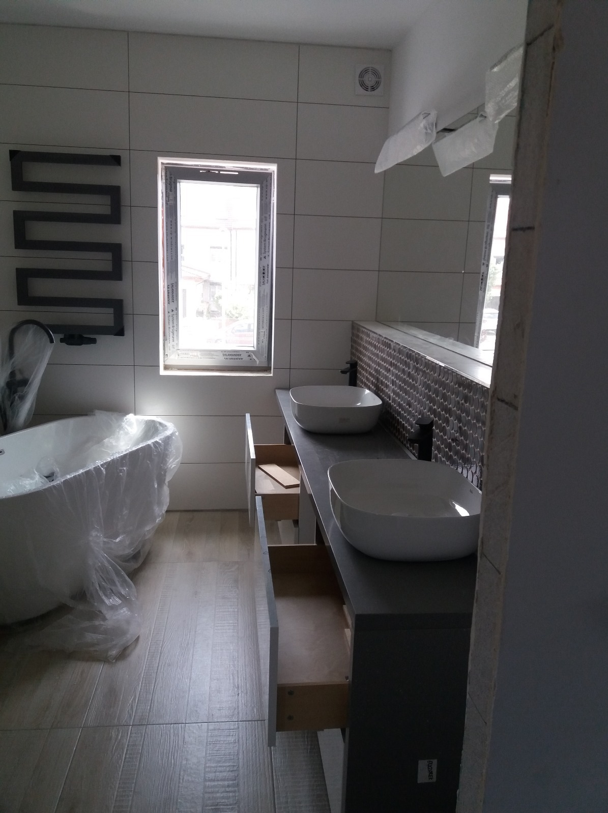 Nowoczesna łazienka w trakcie wykończenia: białe płytki, umywalki nablatowe, otwarta szuflada szafki pod umywalką, wanna w folii ochronnej i grzejnik drabinkowy.