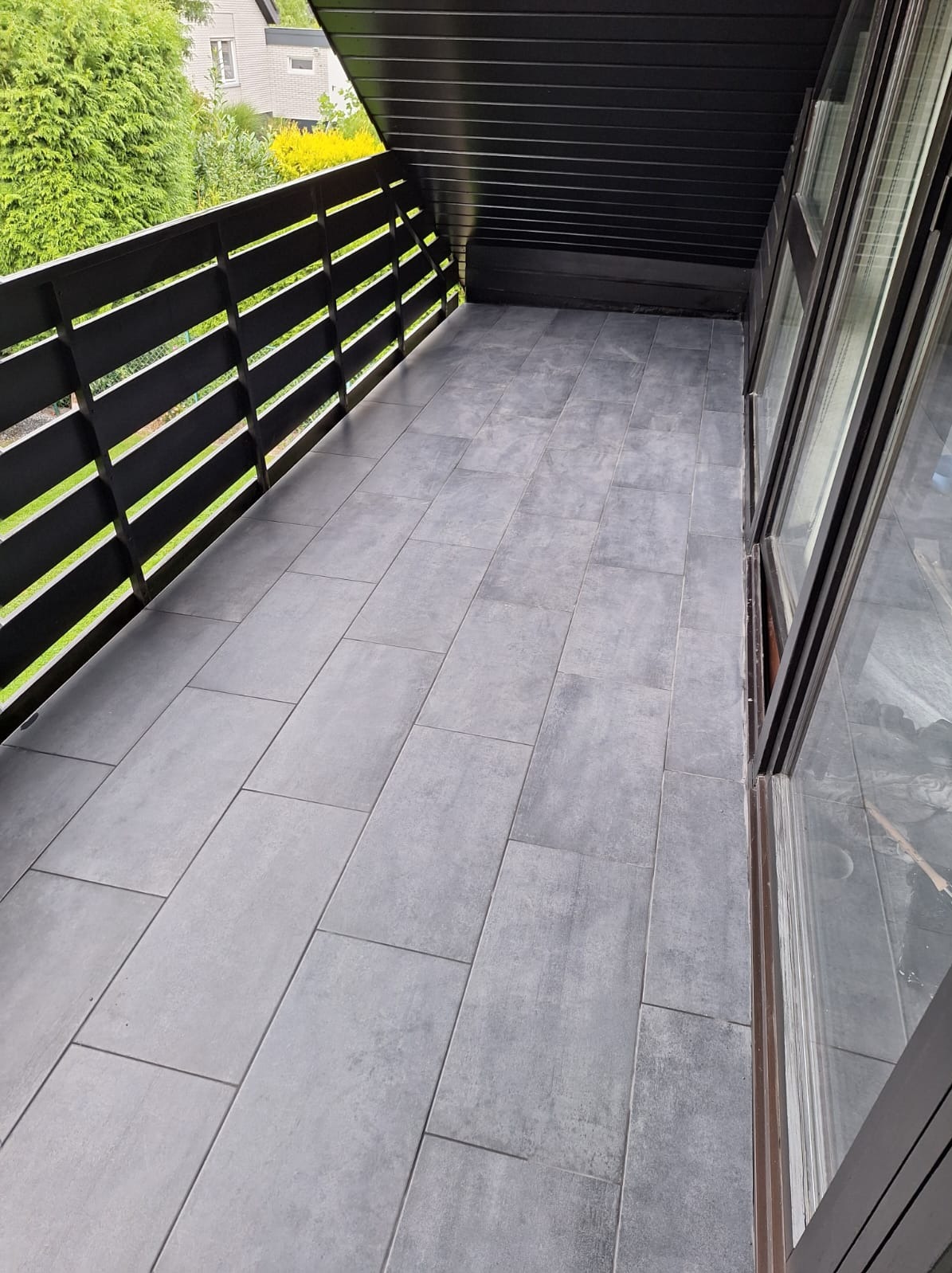 Wyłożony szarymi płytkami balkon z czarną balustradą i stolarką okienną. Widok na zieleń. Układ płytek podłużny, prostopadły do balustrady. Minimalistyczny styl.