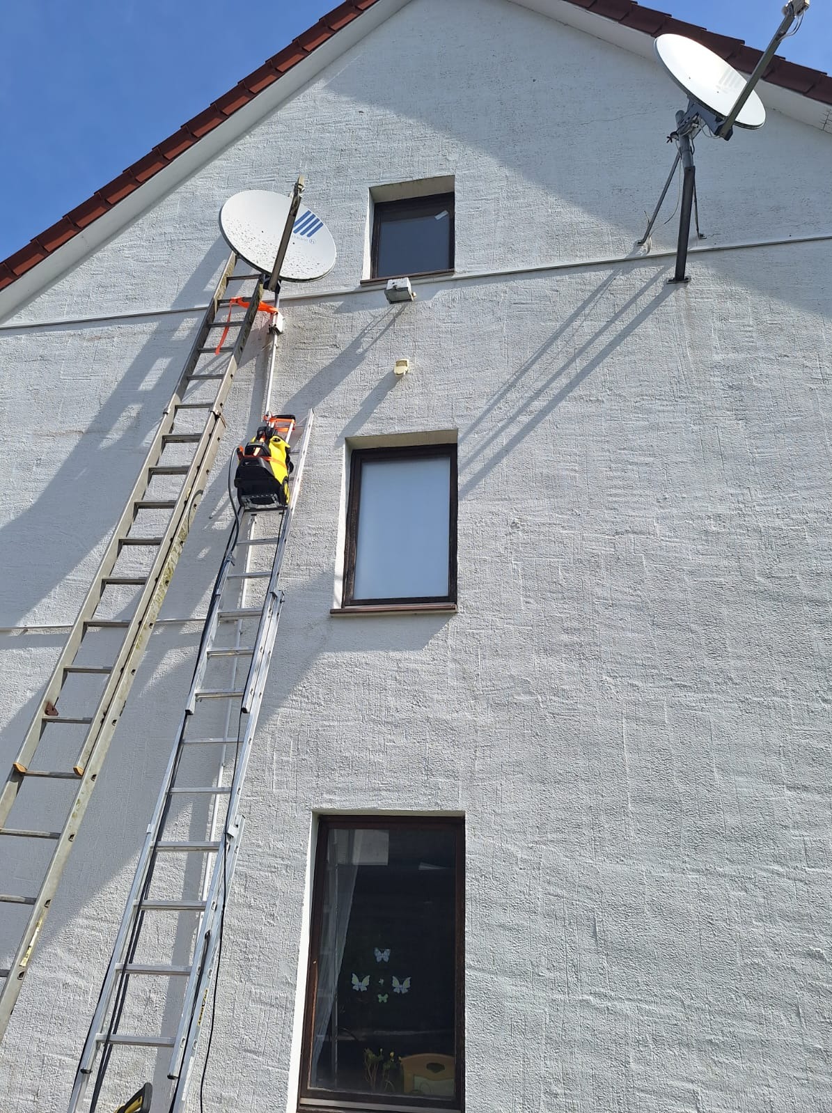 Biała elewacja domu z dwiema antenami satelitarnymi. Do jednej z nich przystawiona drabina z myjką ciśnieniową. Widoczne okna o ciemnych ramach.
