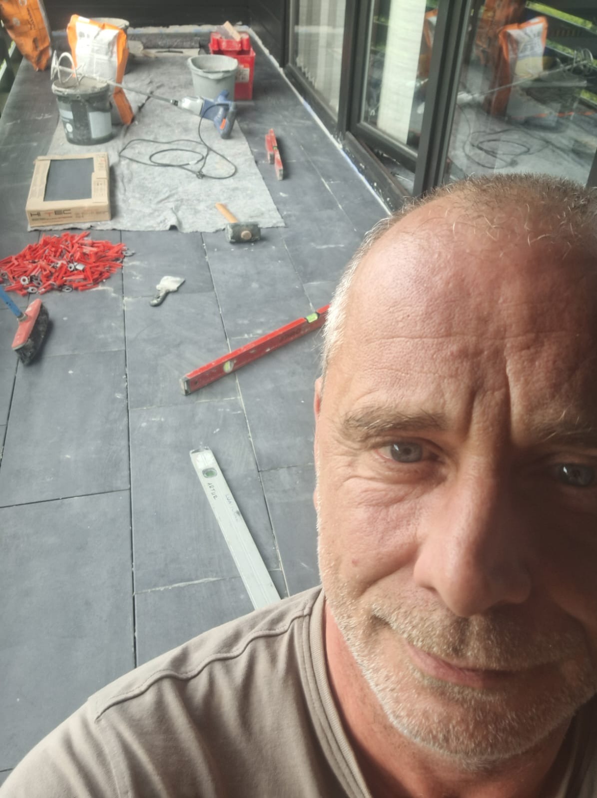 Układanie ciemnych płytek na podłodze balkonu, widoczne narzędzia, poziomica i akcesoria montażowe. W kadrze twarz wykonawcy.
