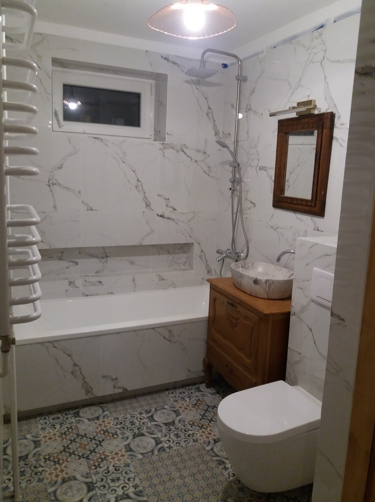 Wykończona łazienka z wanną w zabudowie, umywalką na drewnianej komodzie i toaletą. Białe płytki imitujące marmur i wzorzysta podłoga.