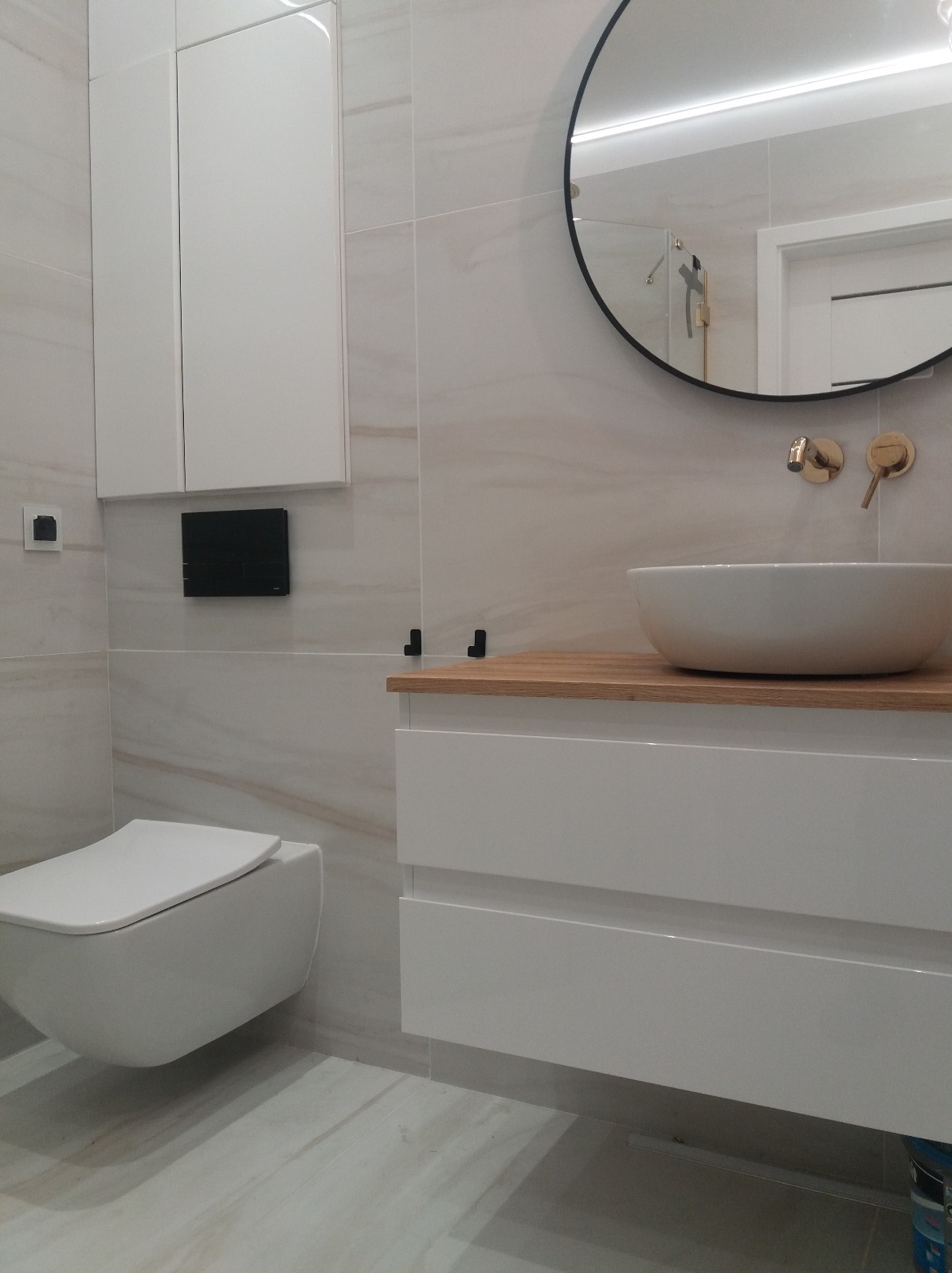 Nowoczesna łazienka z podwieszaną toaletą, umywalką nablatową na drewnianym blacie i okrągłym lustrem z czarną ramą, minimalistyczny design.