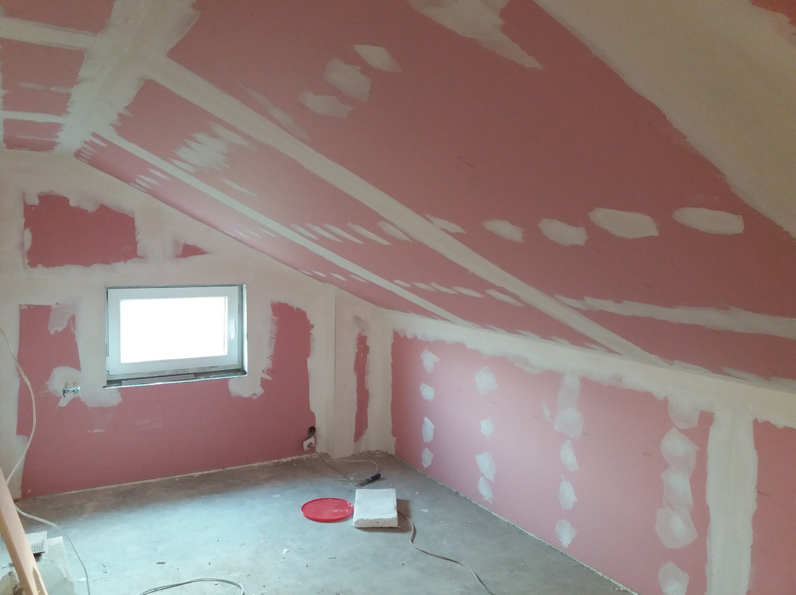 Poddasze w trakcie prac wykończeniowych: różowe płyty gipsowe z widocznymi łączeniami i szpachlowaniem. Okno w ścianie, surowy beton na podłodze, narzędzia pracy.