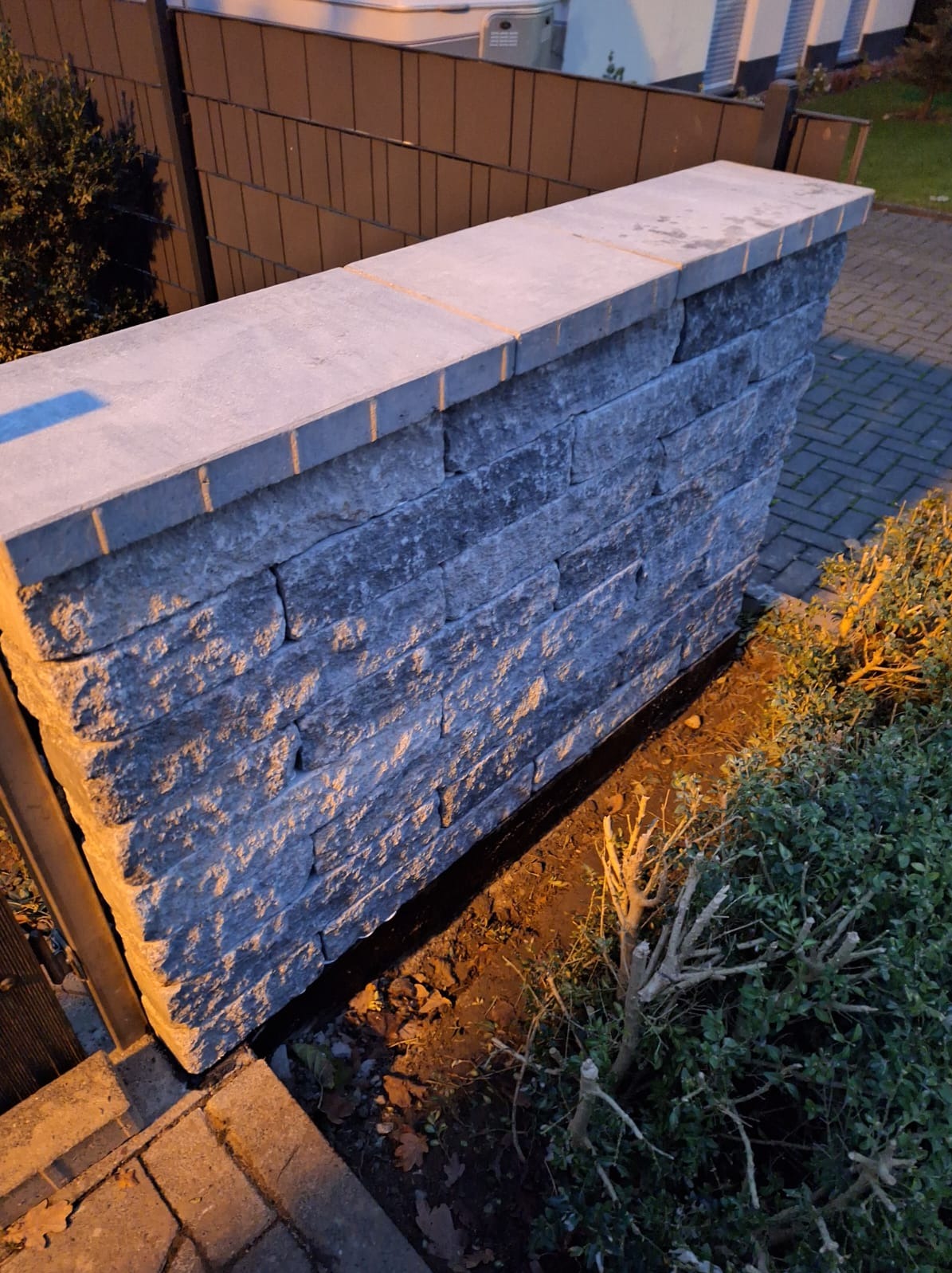 Murowana ścianka z szarego kamienia łupanego, zwieńczona płytami, oddzielająca posesję od chodnika. Widoczne elementy ogrodzenia i zieleń.