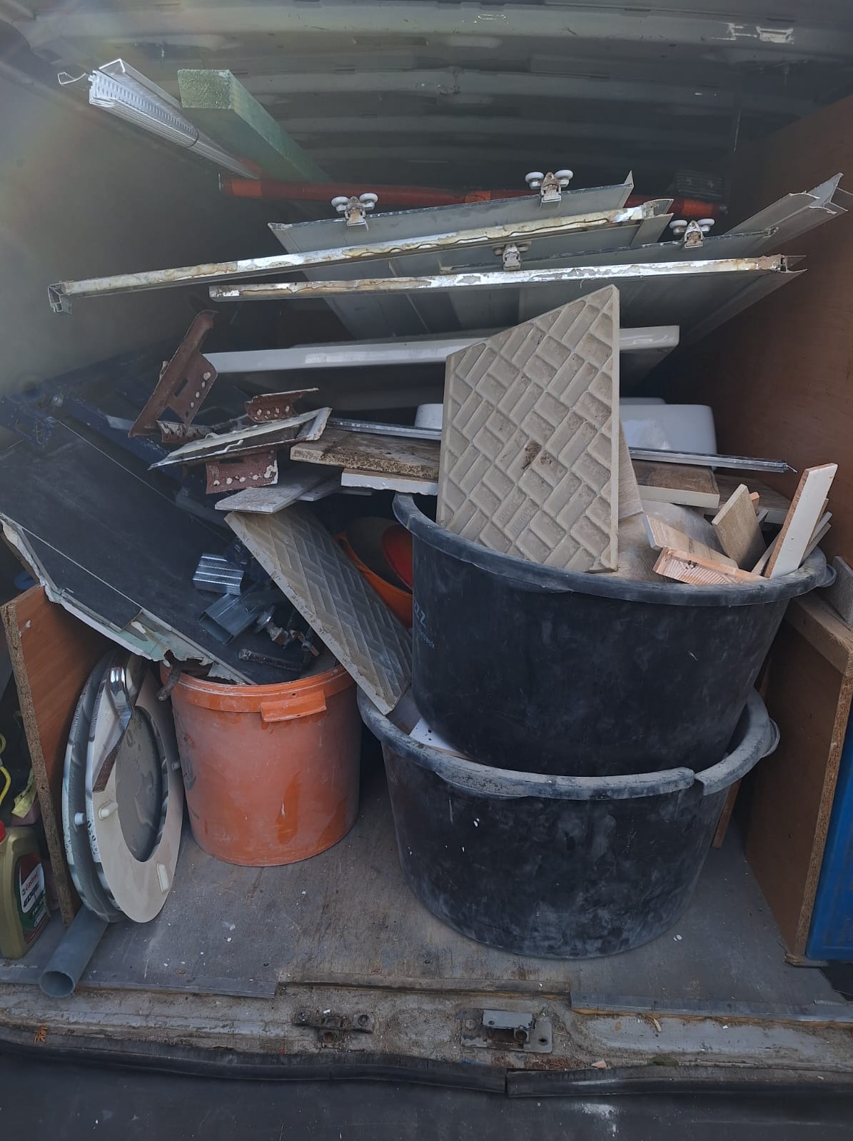 Wnętrze brudnego samochodu dostawczego wypełnione po brzegi materiałami budowlanymi, w tym płytkami, profilami i wiadrami.