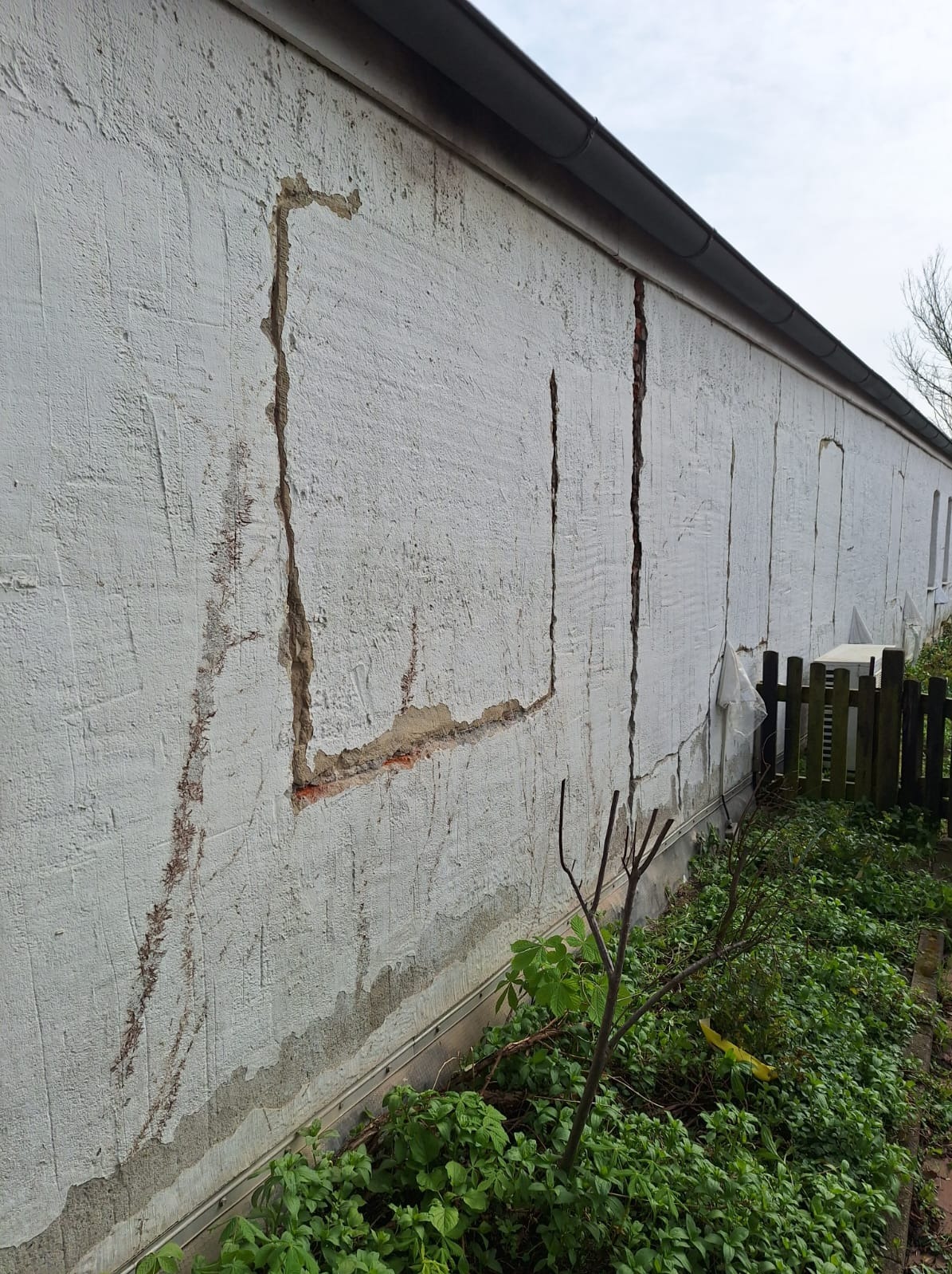 Uszkodzona, biała ściana budynku z widocznymi pęknięciami i ubytkami tynku, z elementami odsłoniętej cegły. Widoczne rośliny u podstawy ściany i fragment ogrodzenia.