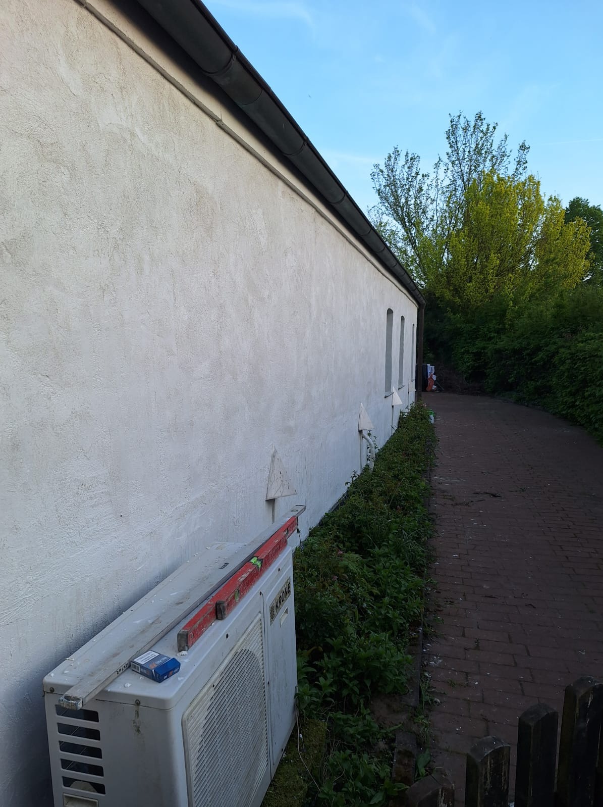 Elewacja budynku z klimatyzatorem i poziomicą, widok z boku. Teksturowana ściana, fragment ścieżki z kostki brukowej, zieleń wzdłuż ściany, błękitne niebo.