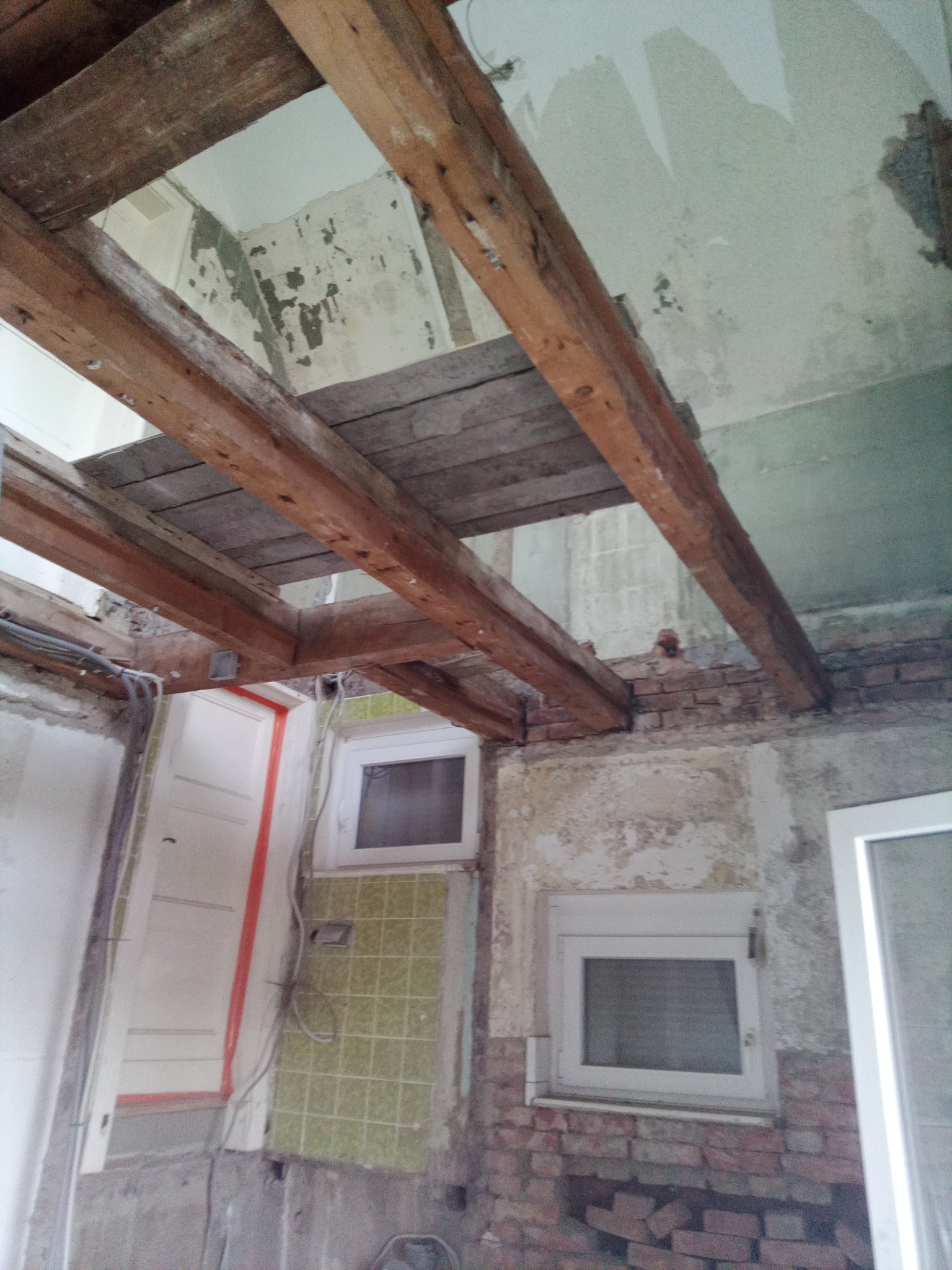 Klatka schodowa w trakcie remontu: drewniane belki stropowe, odsłonięte cegły i resztki starej glazury. Widoczne okna i instalacje elektryczne.