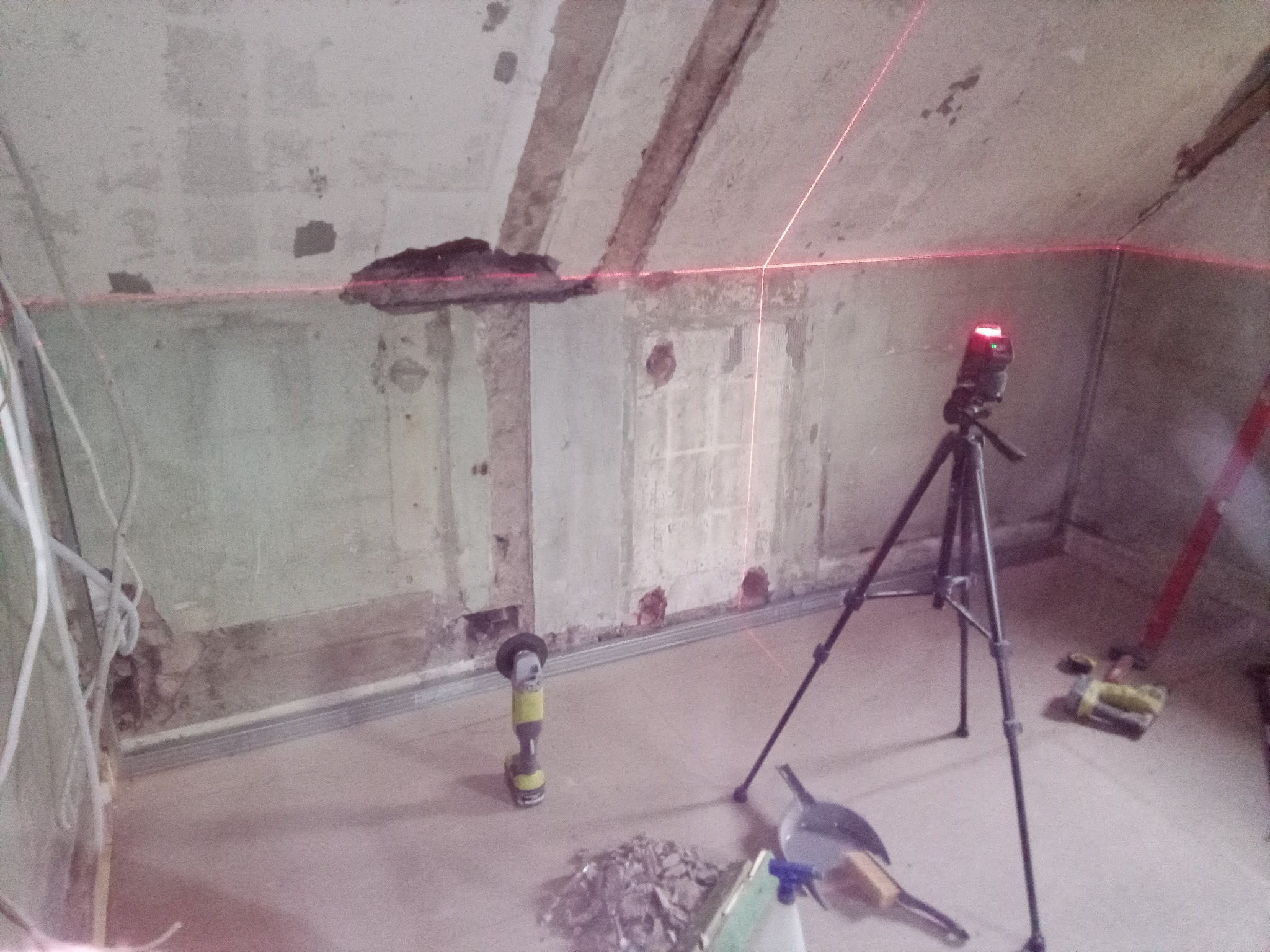 Remont poddasza: laser krzyżowy wyznacza poziom na ścianie z odsłoniętym murem, obok szlifierka kątowa i gruz. Etap przygotowawczy przed wykończeniem.