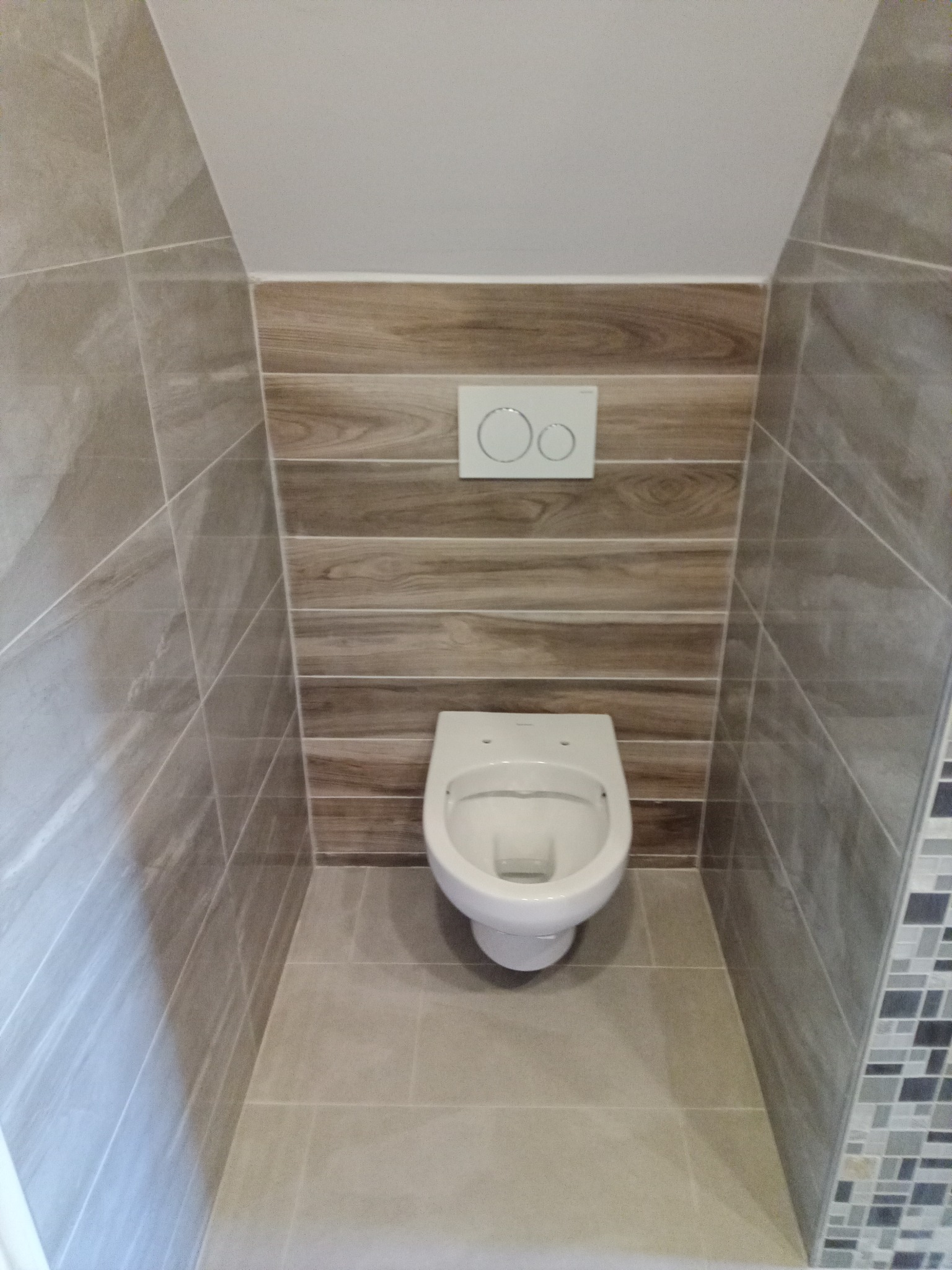 Kompaktowa toaleta w beżowo-brązowej łazience z drewnopodobnymi płytkami na ścianie i mozaiką. Ukończona aranżacja małej przestrzeni z wiszącą miską WC.