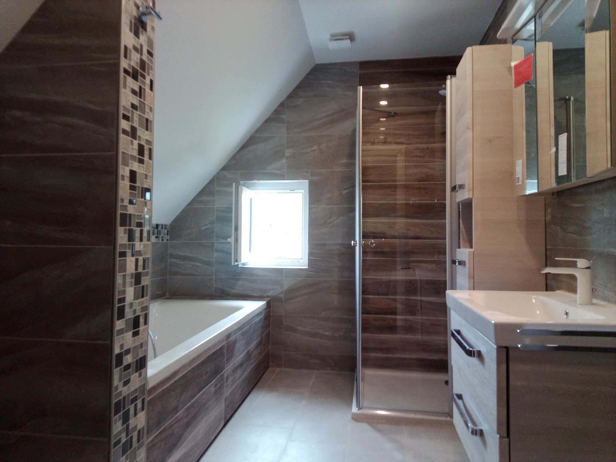 Nowoczesna łazienka na poddaszu z wanną, kabiną prysznicową i umywalką w odcieniach szarości i brązu. Uwagę zwracają mozaiki na obudowie wanny.