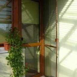 Adrian Kowalczyk - Otwarta moskitiera ramkowa w drewnianej oprawie, z widoczną siatką i zawiasami, zamontowana w drzwiach balkonowych. Obok doniczka z rośliną.