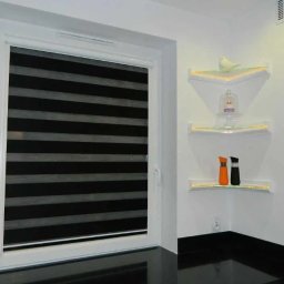 Adrian Kowalczyk - Okno z roletą dzień noc w czarno-białe pasy. Na ścianie trzy szklane półki narożne z oświetleniem LED, na których stoją dekoracje.