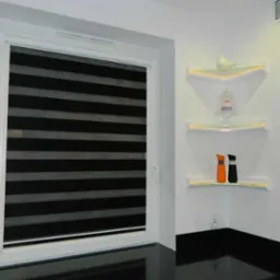 Okno z roletą dzień noc w czarno-białe pasy. Na ścianie trzy szklane półki narożne z oświetleniem LED, na których stoją dekoracje.