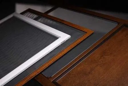 Różne ramki moskitier: biała aluminiowa, brązowa drewnopodobna i szara, ułożone warstwowo, ukazujące detale siatki i wykończenia. Ciemne tło.