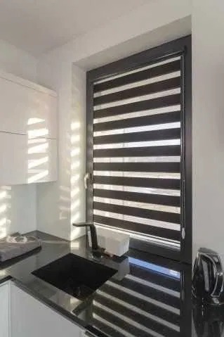 Roleta dzień noc w czarnej ramie okiennej, zamontowana w kuchni z czarnym blatem. Widoczne refleksy światła na blacie i szafce.