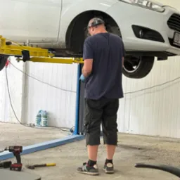 Mechanik w warsztacie, w niebieskiej koszulce, pracuje przy podniesionym na podnośniku białym samochodzie. Widoczne narzędzia i butelki z wodą w tle.