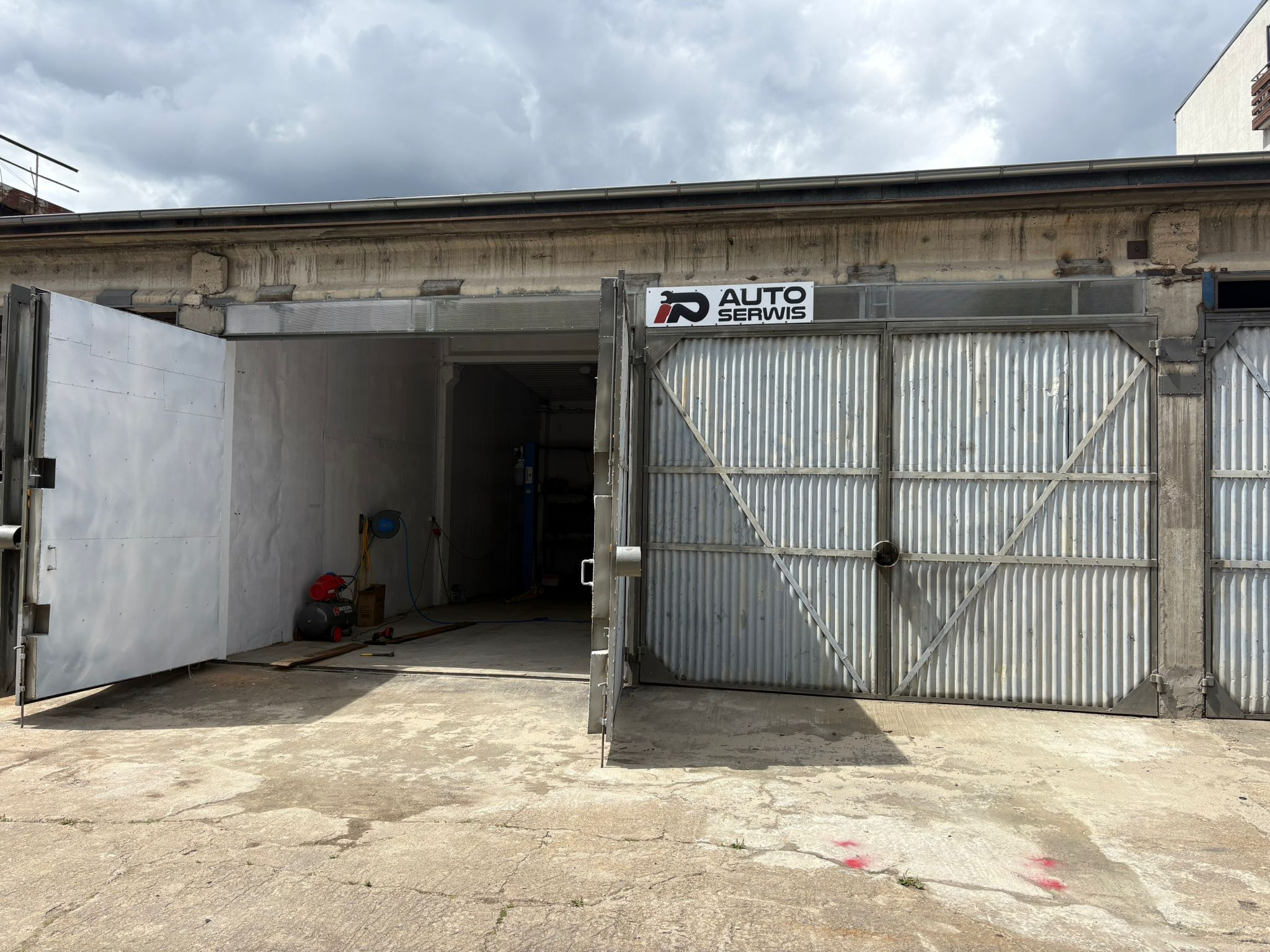 Otwarta brama warsztatu samochodowego z widocznym podnośnikiem wewnątrz, betonowa posadzka, znak 'Auto Serwis' nad bramą, pochmurne niebo w tle.