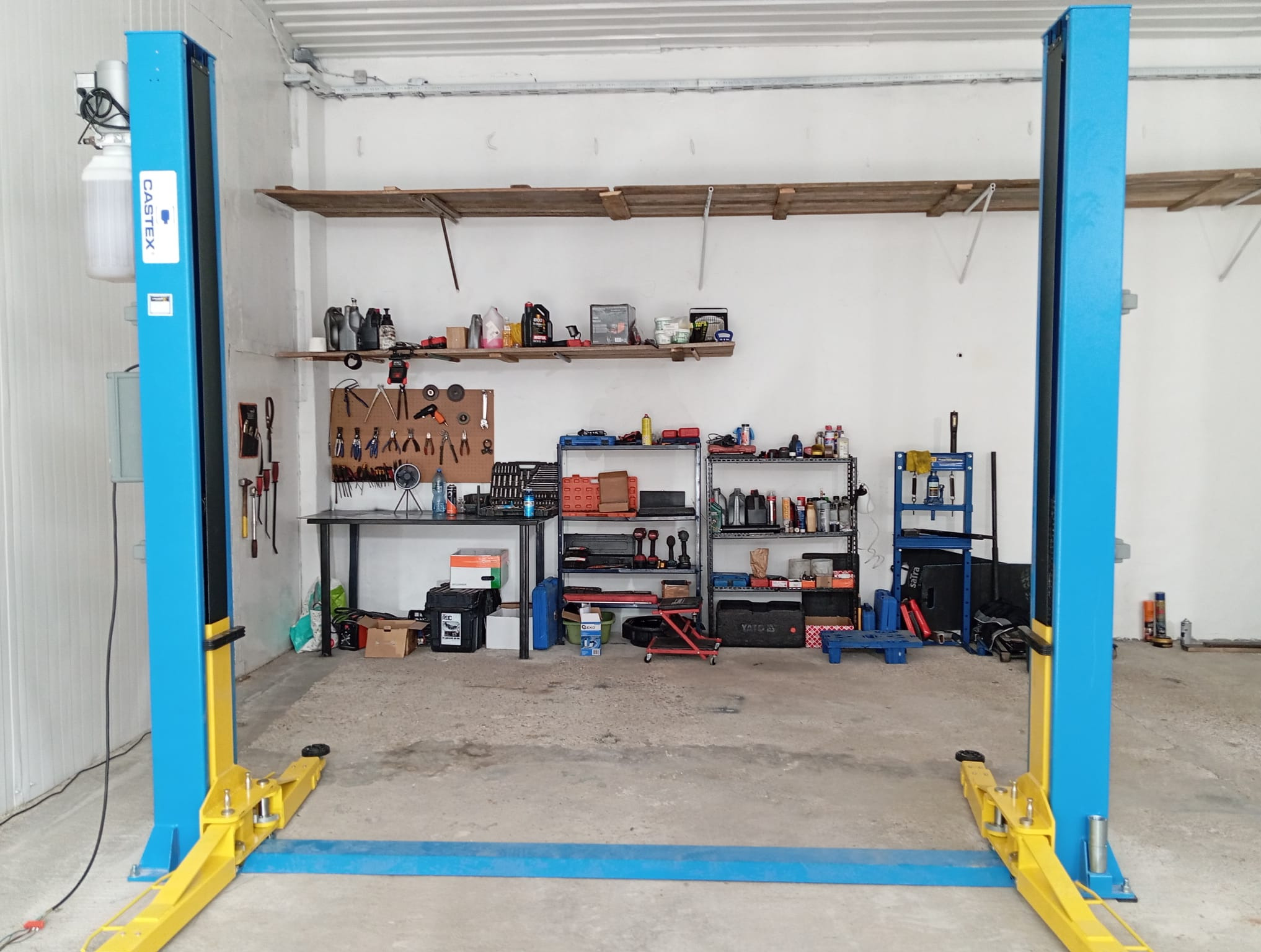 Dwukolumnowy podnośnik samochodowy Castex w warsztacie, z narzędziami i olejami na półkach w tle. Niebiesko-żółta konstrukcja na betonowej posadzce.