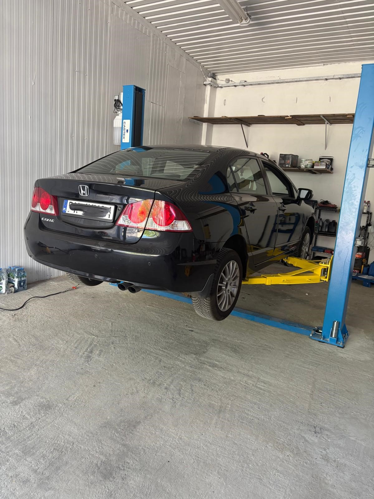 Czarny samochód Honda Civic na podnośniku w warsztacie. Widoczne tylne światła, koła i podwozie. W tle regały z narzędziami i akcesoriami.