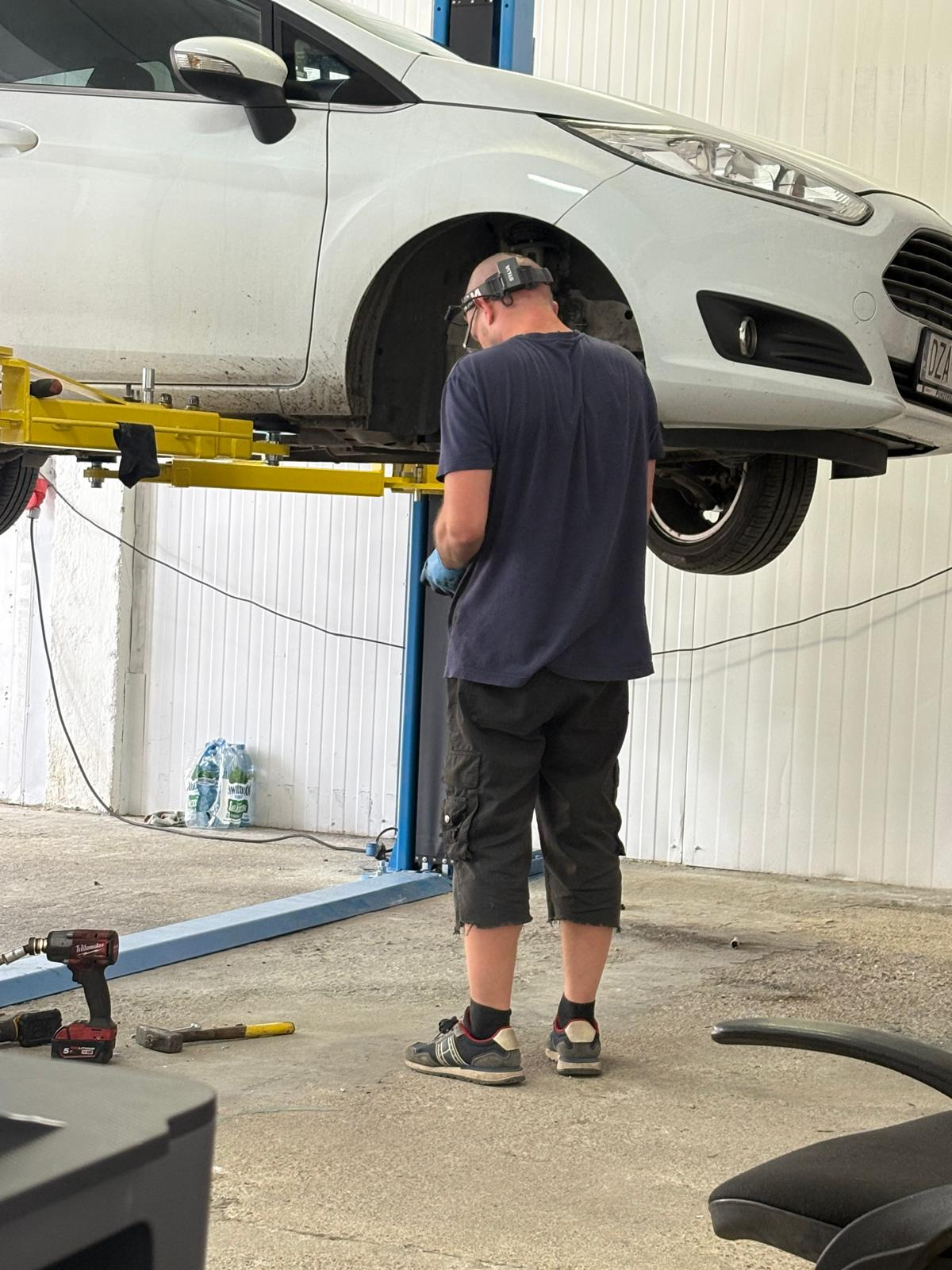 Mechanik w warsztacie, w niebieskiej koszulce, pracuje przy podniesionym na podnośniku białym samochodzie. Widoczne narzędzia i butelki z wodą w tle.