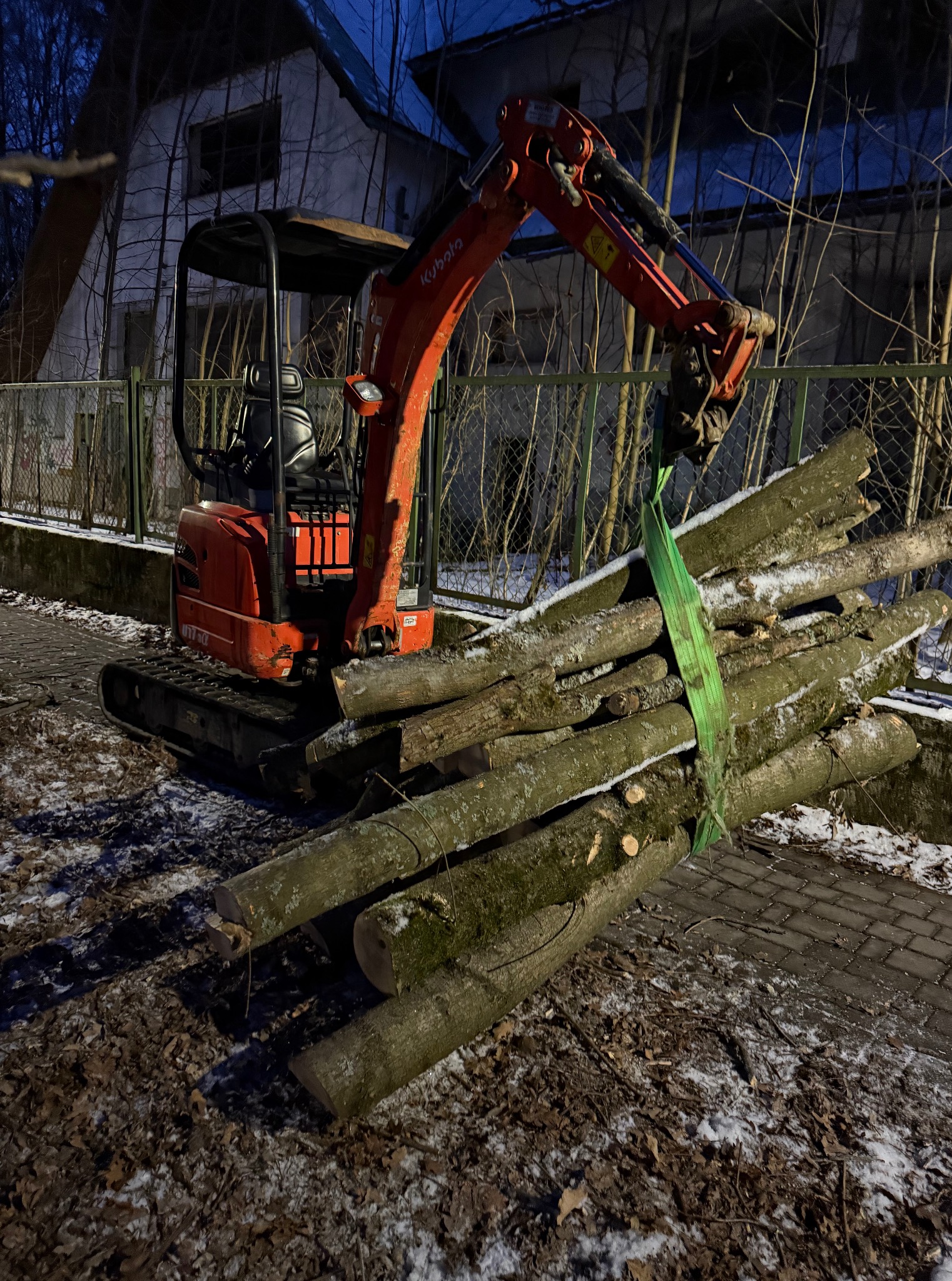 Pomarańczowa mini koparka Kubota podnosi pęk ściętych pni drzew za pomocą zielonego pasa. W tle budynek i drzewa.