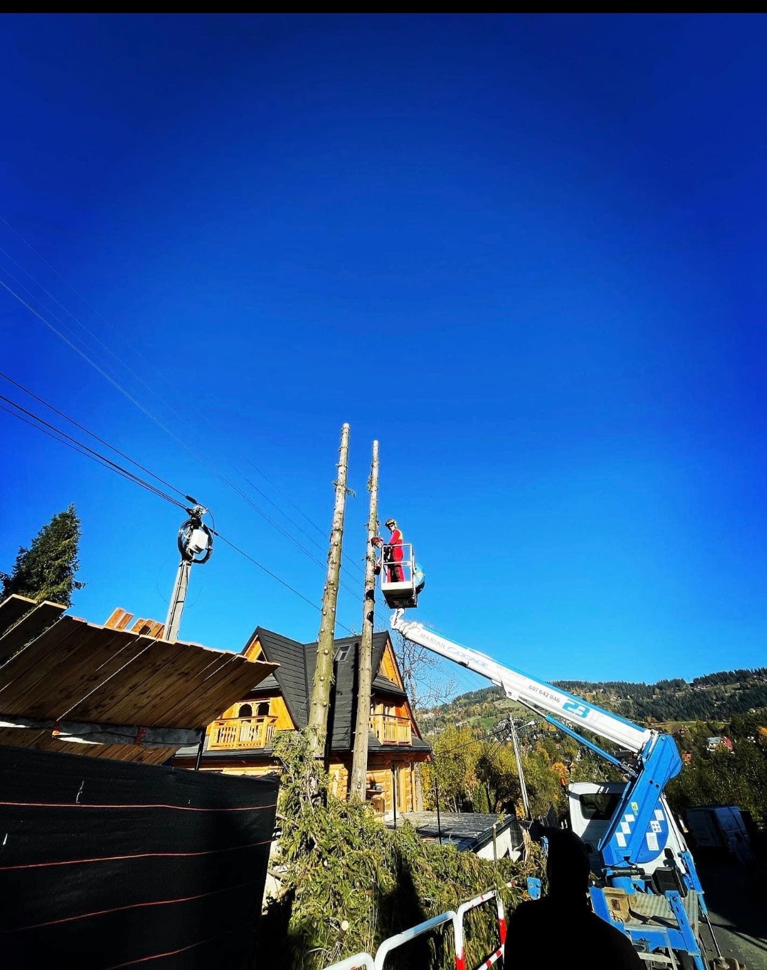 Pracownik w czerwonym kombinezonie na podnośniku przycina wysokie drzewo obok drewnianego domu. W tle góry i błękitne niebo.