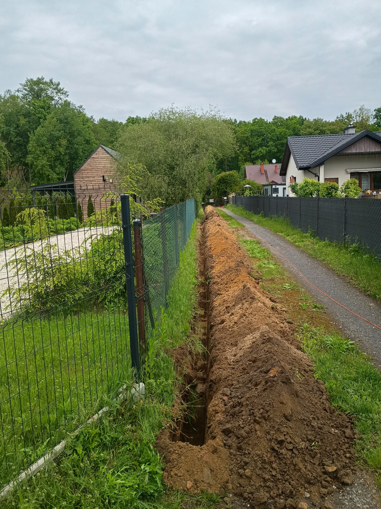 Wykopany rów wzdłuż ogrodzenia na posesji, prawdopodobnie pod kable telekomunikacyjne. Widoczny kabel leżący obok ścieżki. W tle domy i zieleń.