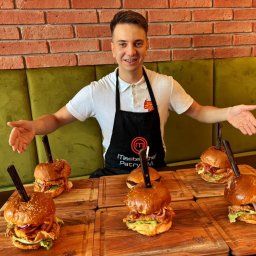 TUR-RESTAURACJA Bartosz Zapiec - Uśmiechnięty kucharz prezentuje sześć burgerów na drewnianych deskach, każdy z nożem. W tle ceglana ściana i zielona kanapa.