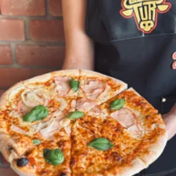 Pizza z szynką, cebulą i bazylią, serwowana na tle ceglanej ściany przez kelnera w Turznie, z logo restauracji na fartuchu. Apetyczny kadr.