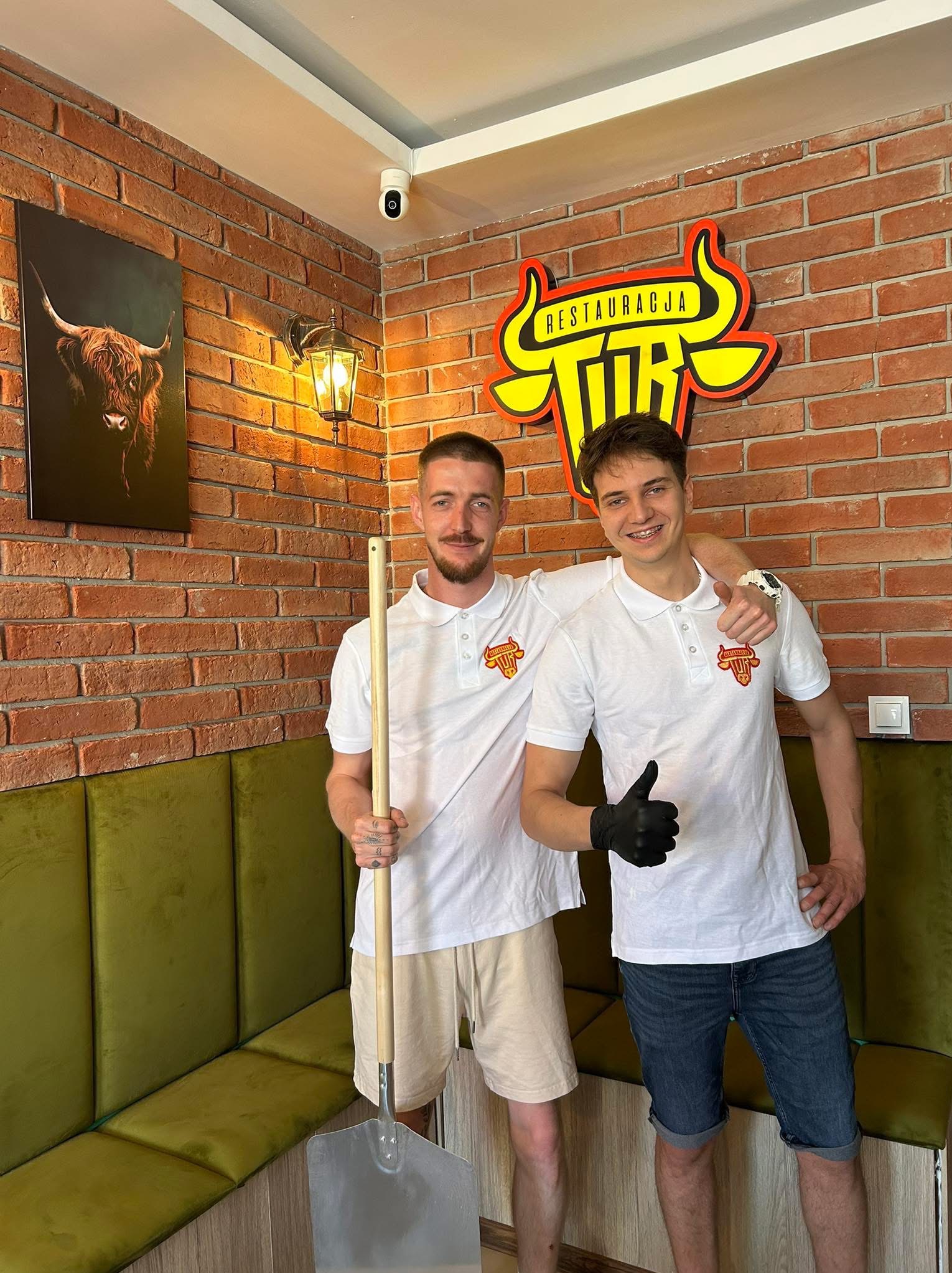Dwóch uśmiechniętych mężczyzn w restauracji 'TUR', jeden trzyma łopatę do pizzy, drugi kciuk w górę. Ceglana ściana z logo i obrazem byka.