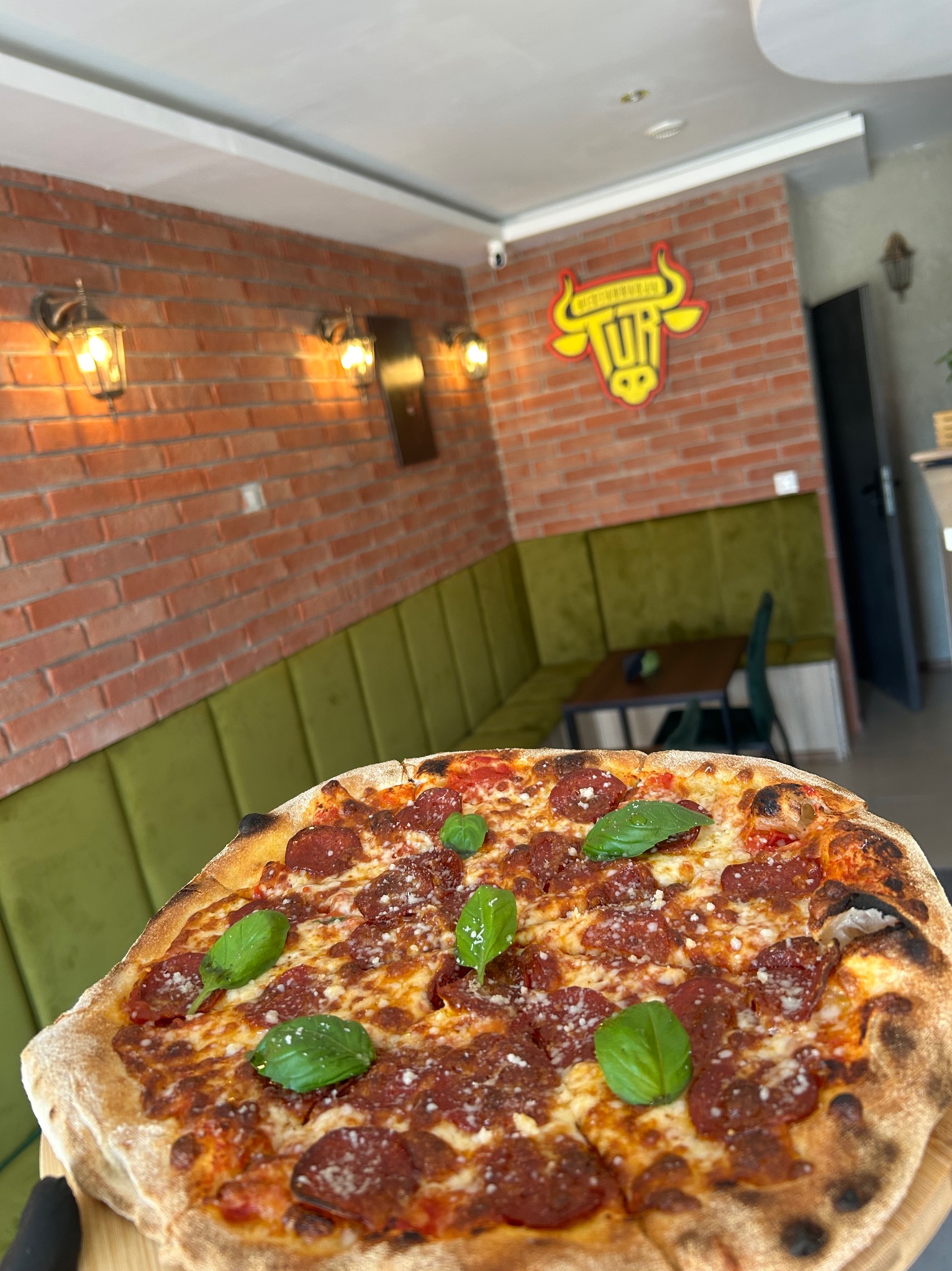 Apetyczna pizza pepperoni z bazylią na tle ceglanej ściany z logo i zieloną kanapą, prezentowana w restauracji. Idealna na firmowy lunch lub świąteczny poczęstunek.