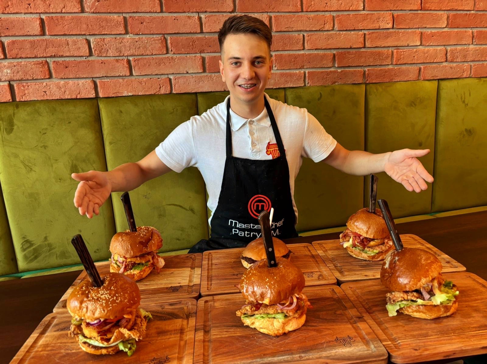 Uśmiechnięty kucharz prezentuje sześć burgerów na drewnianych deskach, każdy z nożem. W tle ceglana ściana i zielona kanapa.