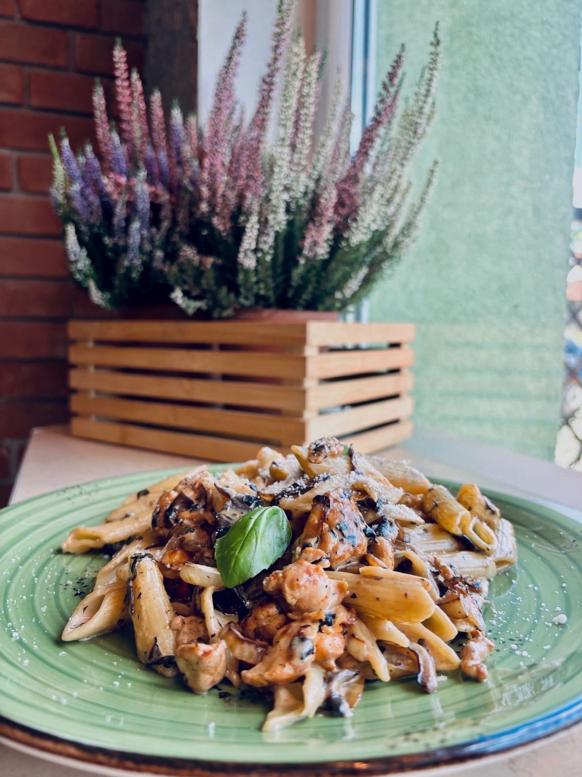 Penne z kurczakiem i grzybami na zielonym talerzu, dekoracja z bazylii. W tle drewniana skrzynka z wrzosem, ceglana ściana i okno.