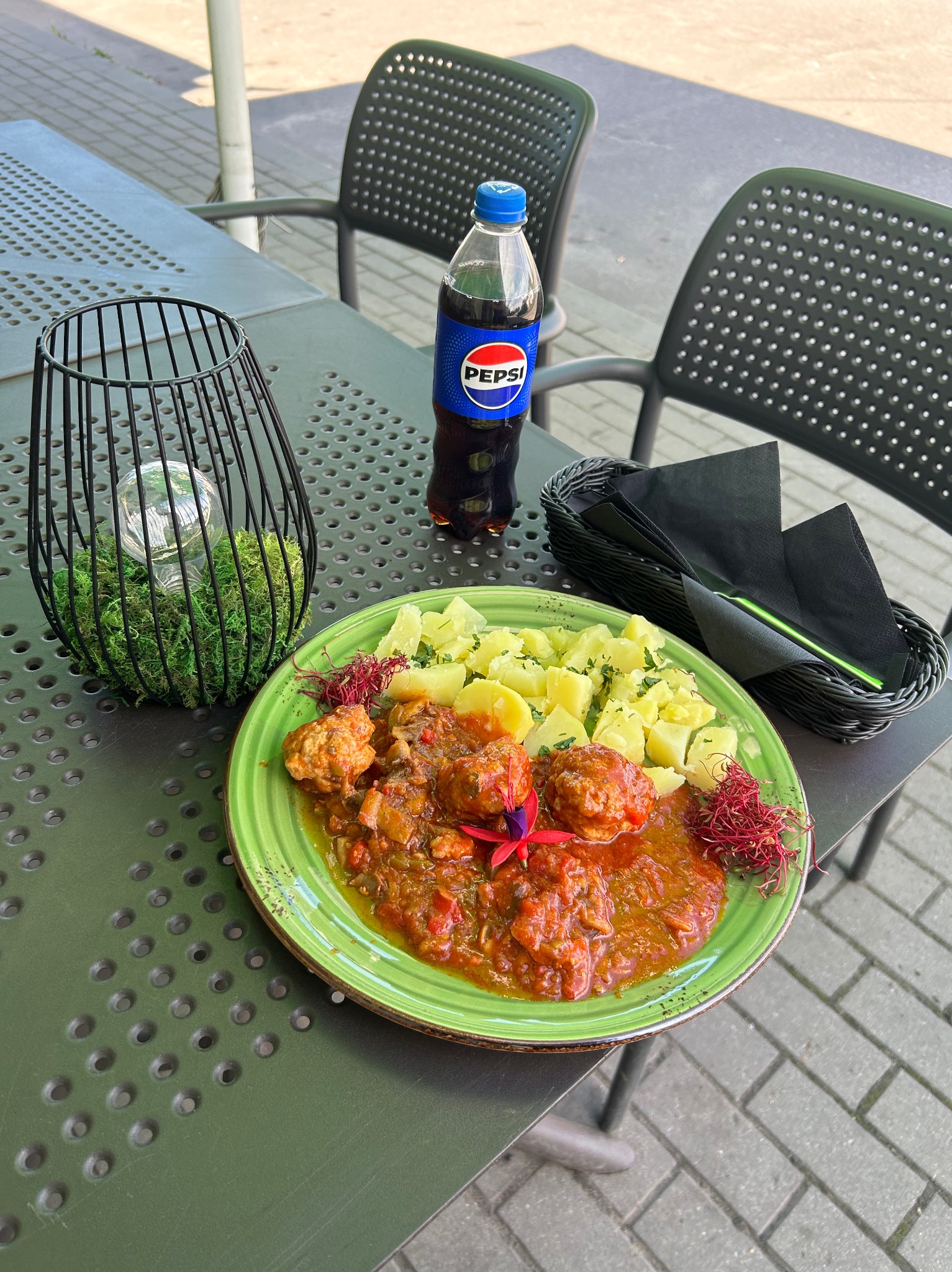Danie z klopsikami w sosie, ziemniakami i dekoracją na zielonym talerzu, obok napój Pepsi, czarne serwetki i dekoracja stołu na zewnątrz.