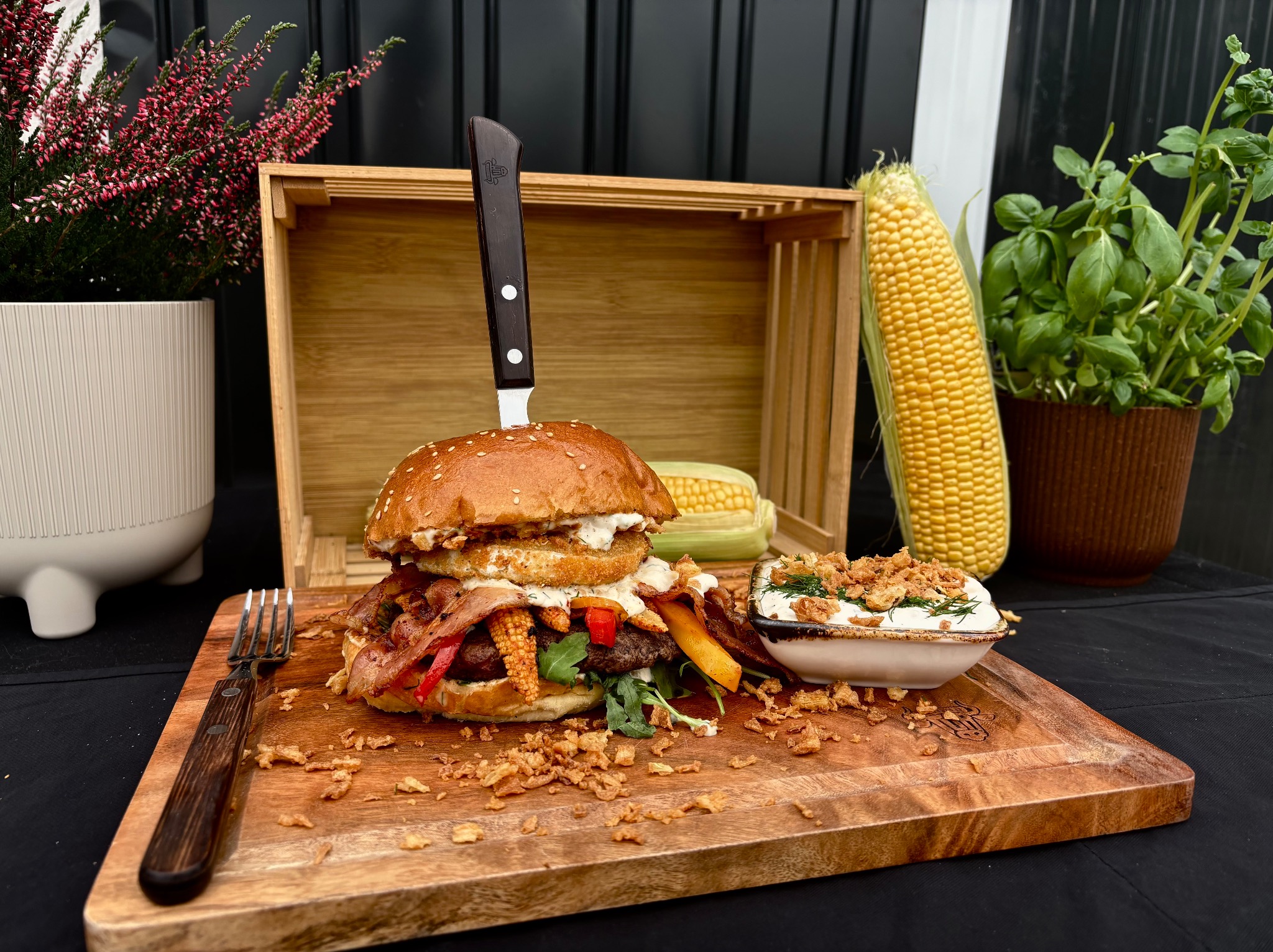 Apetyczny burger z kukurydzą i boczkiem, wbity nożem, na drewnianej desce z sosem, w otoczeniu roślin i skrzynki. Idealny zestaw cateringowy.